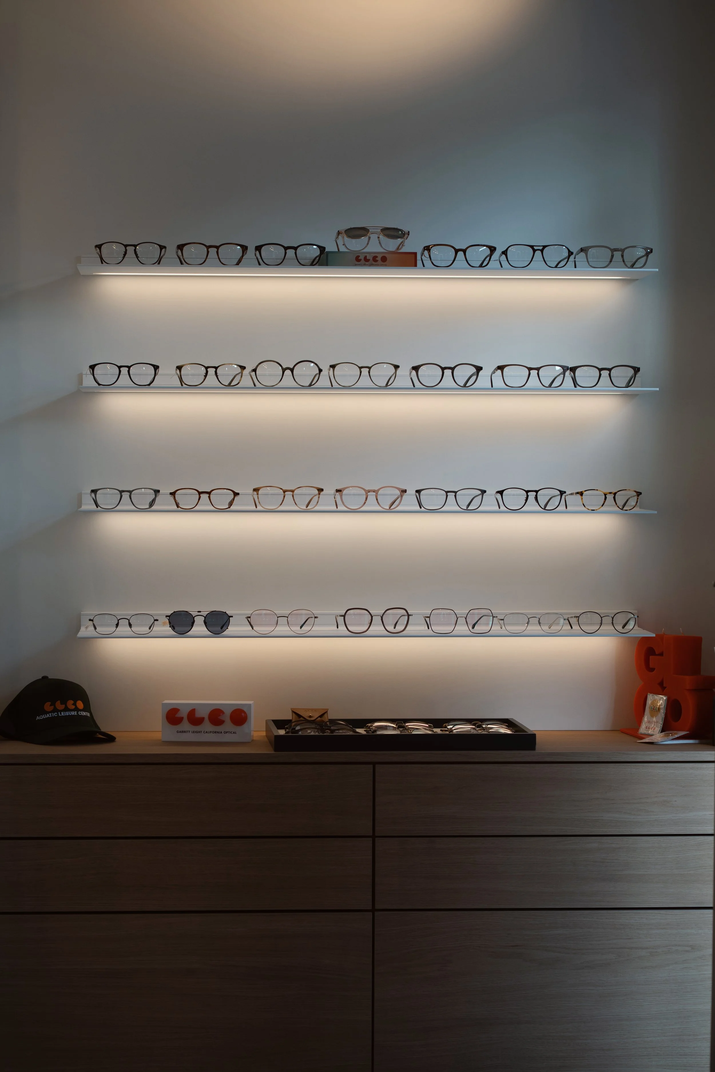 Verlichte wand met meerdere planken vol brilmonturen in een optiekzaak, inclusief een display van Garrett Leight California Optical.