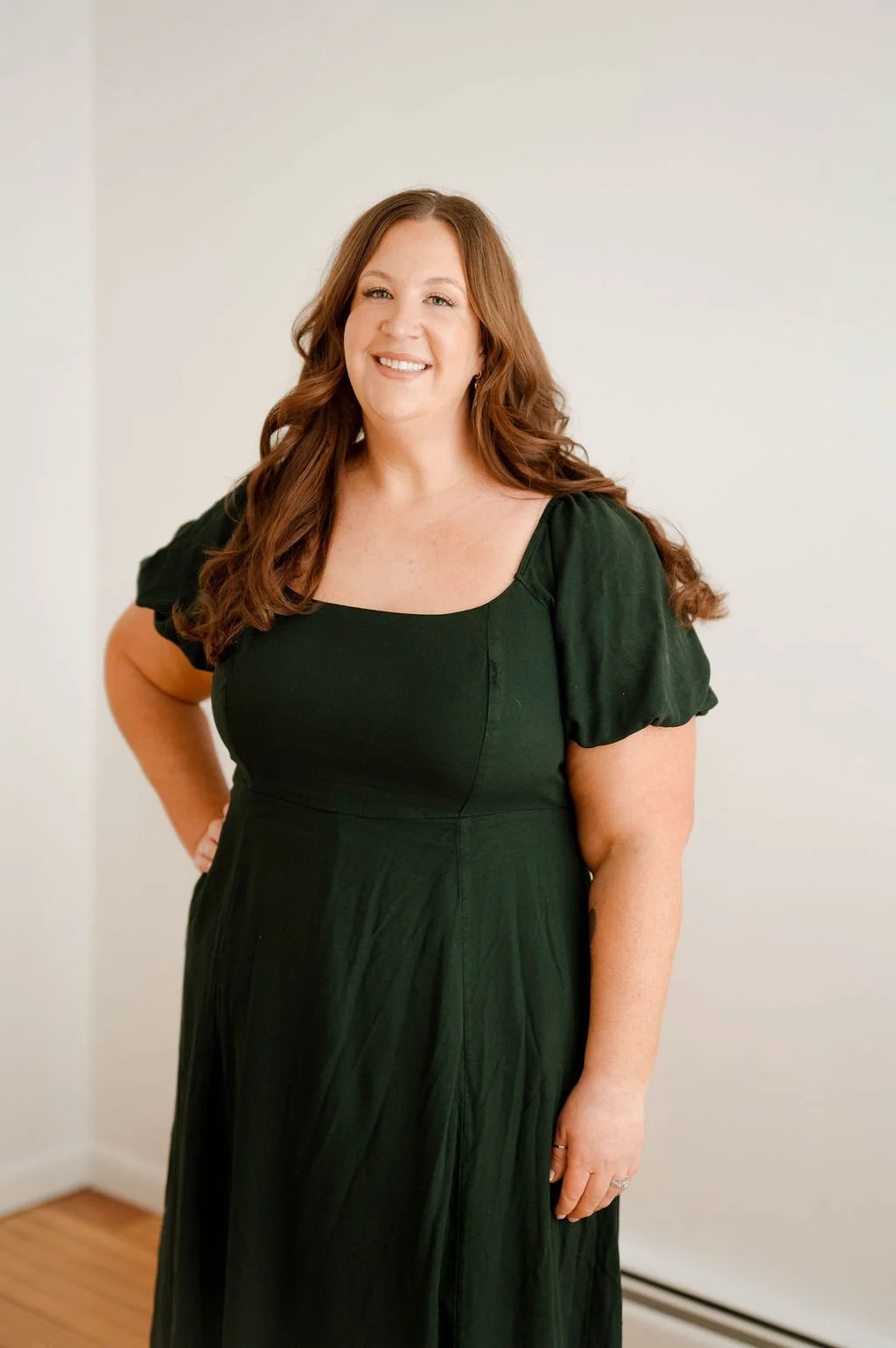 A woman with long, wavy, auburn hair, smiling, wearing a dark green dress with puffed short sleeves, standing in a minimalist room with a plain off-white wall background.
