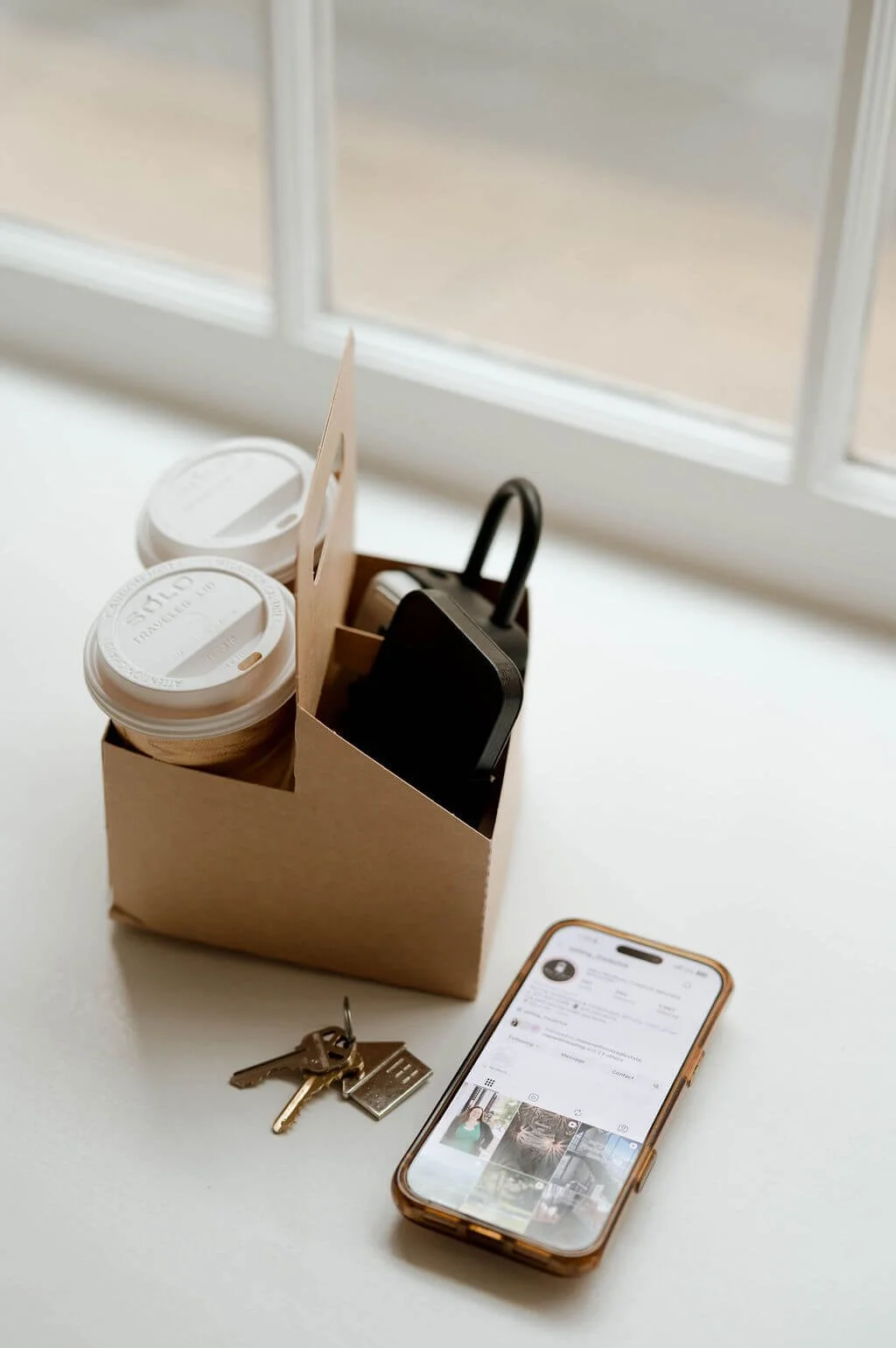 Coffee cups, house keys, and a phone on a table by a sunny window