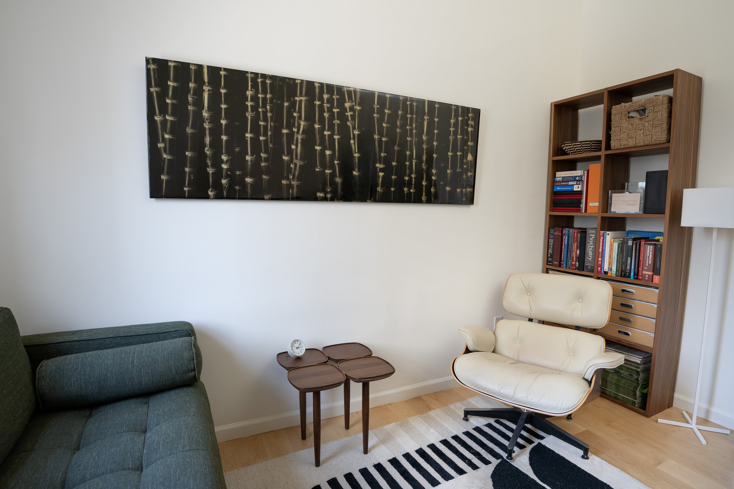 A cozy therapy office with a green sofa, a beige mid-century modern chair, a small wooden side table, a black and white striped rug, a wooden bookshelf filled with books, a large abstract wall art, and a floor lamp with a white shade.