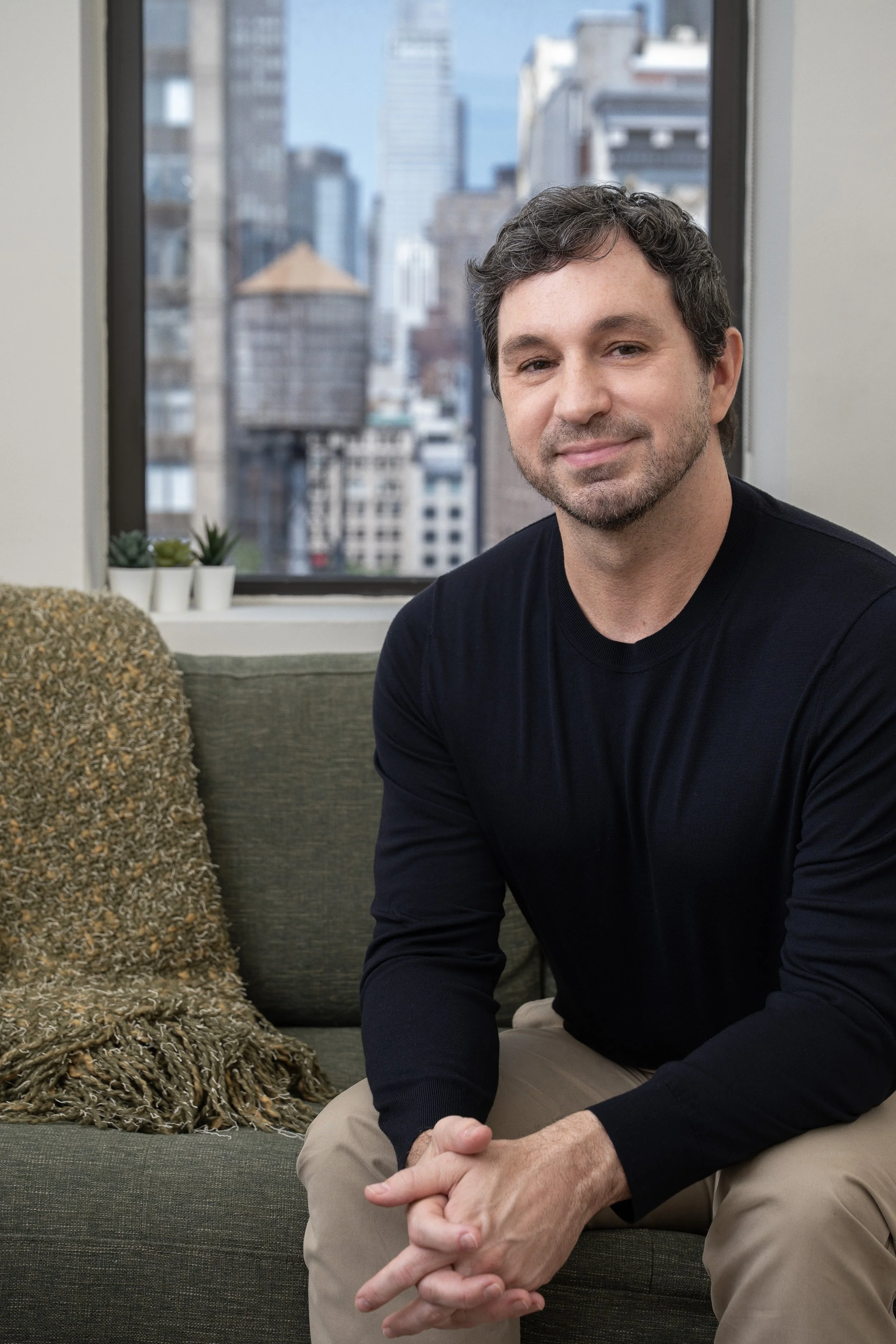 A man with dark curly hair and a beard, wearing a navy sweater, sitting on a couch in front of a window with a cityscape background, smiling softly.