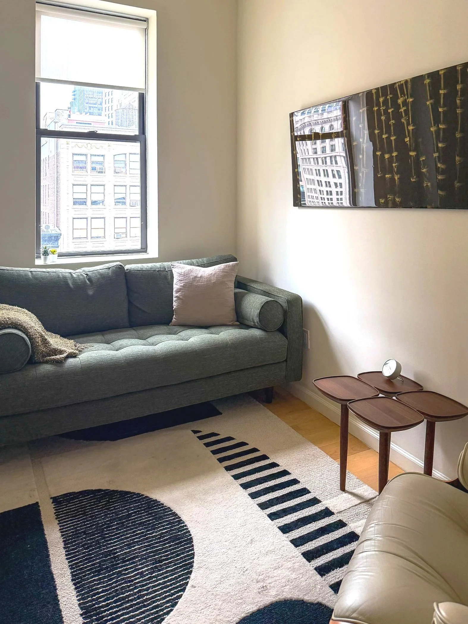 Living room with a green sofa, a beige armchair, a rug with black and white patterns, a wall mirror, a window with city buildings view, and a small side table with a clock.
