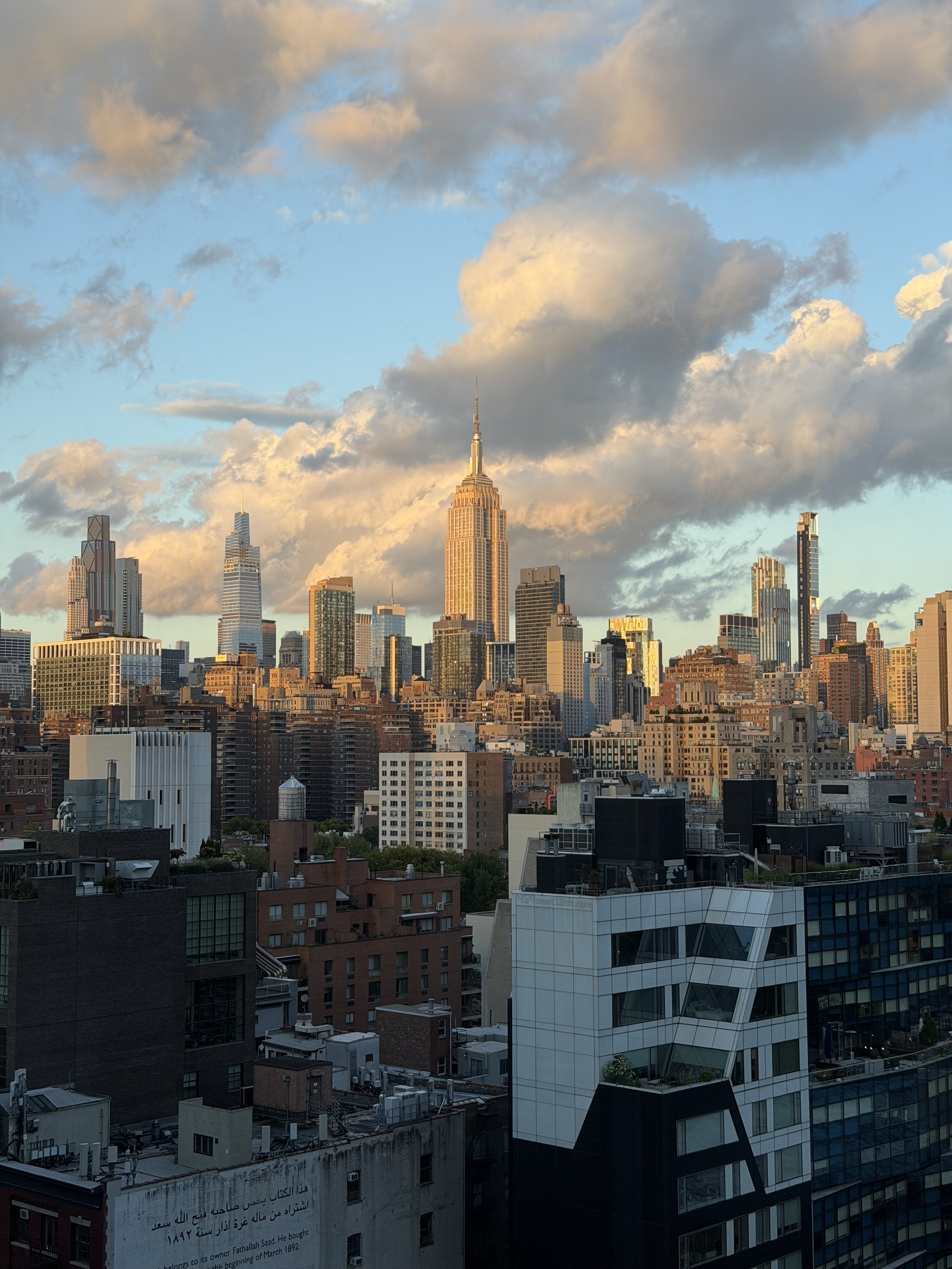 A cityscape of skyscrapers during sunset with a partly cloudy sky in the background. The Empire State Building is prominently visible in the center.