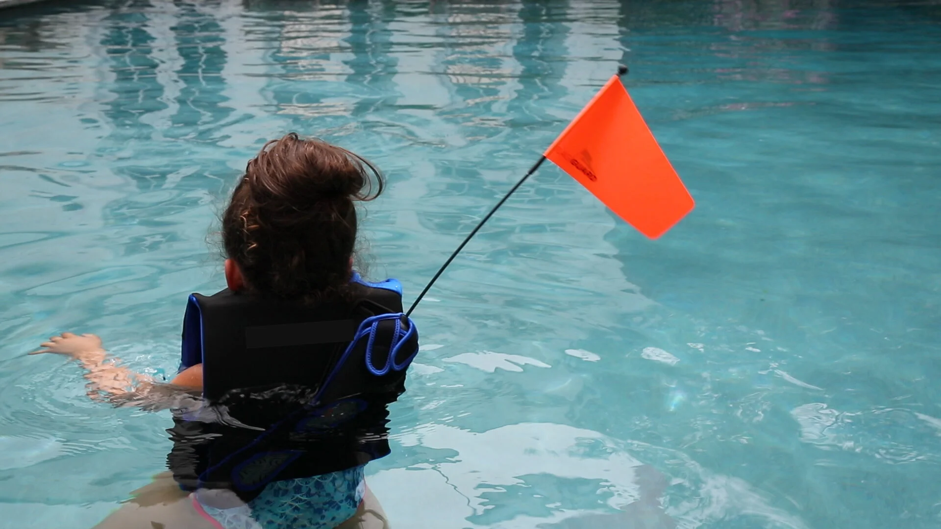 Child in a life jacket floating in the water, holding a small orange safety flag in a pool.
