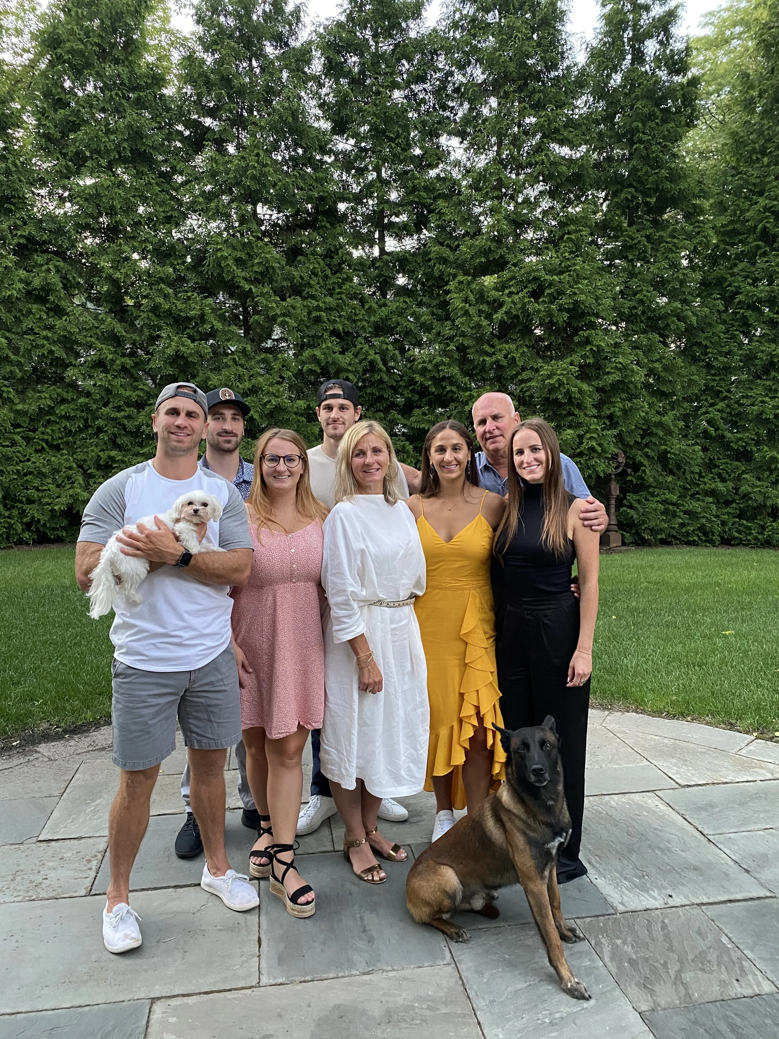 A group of nine people and two dogs standing outdoors on a paved area in front of a large green tree and a grassy lawn.