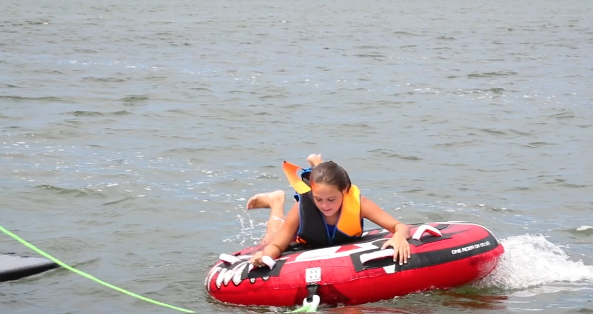 A young girl in a life jacket is falling off a red and black inflatable watercraft into the water, with a lake or ocean in the background.