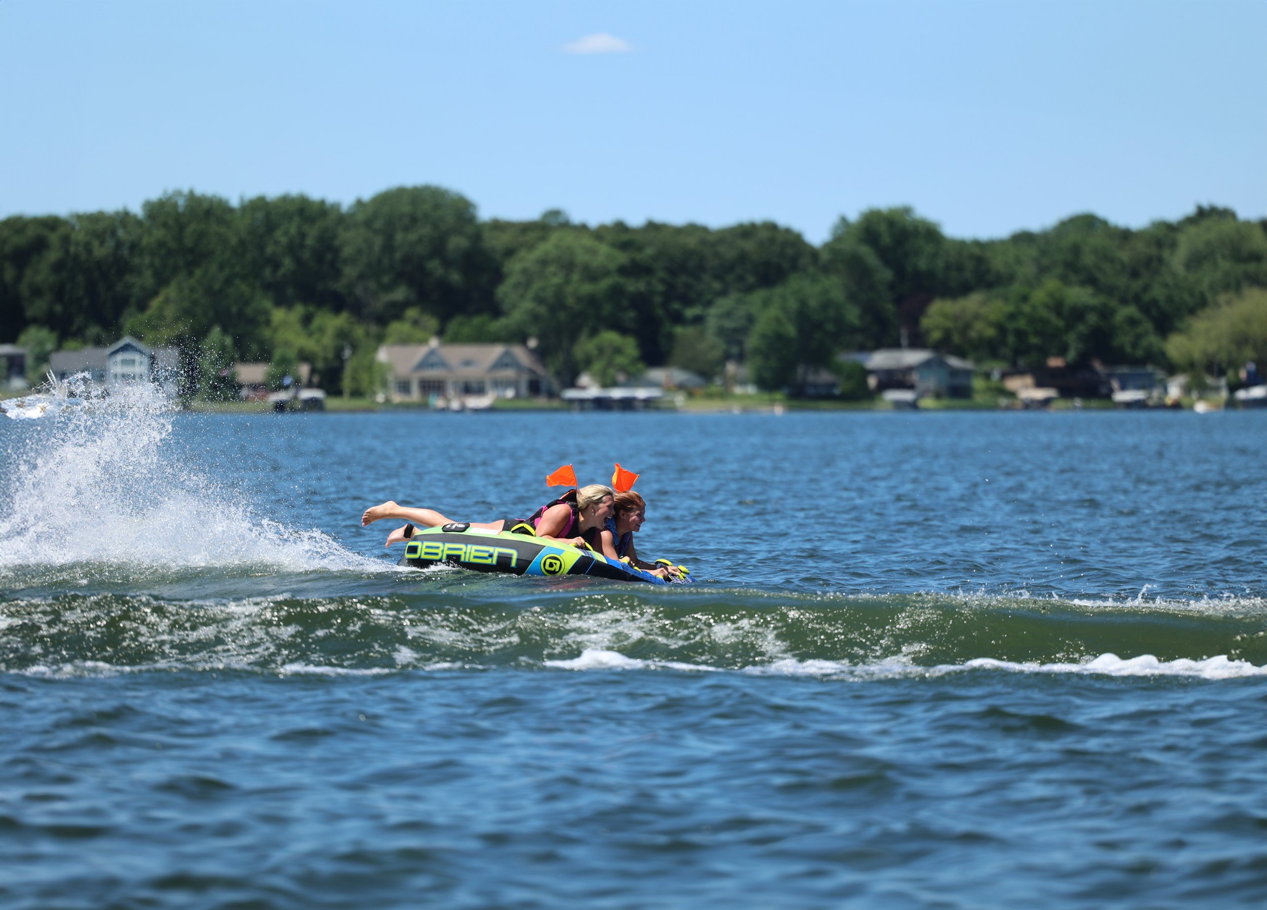 Two people riding a jet ski on a lake with houses and a wooded shoreline in the background under a blue sky.
