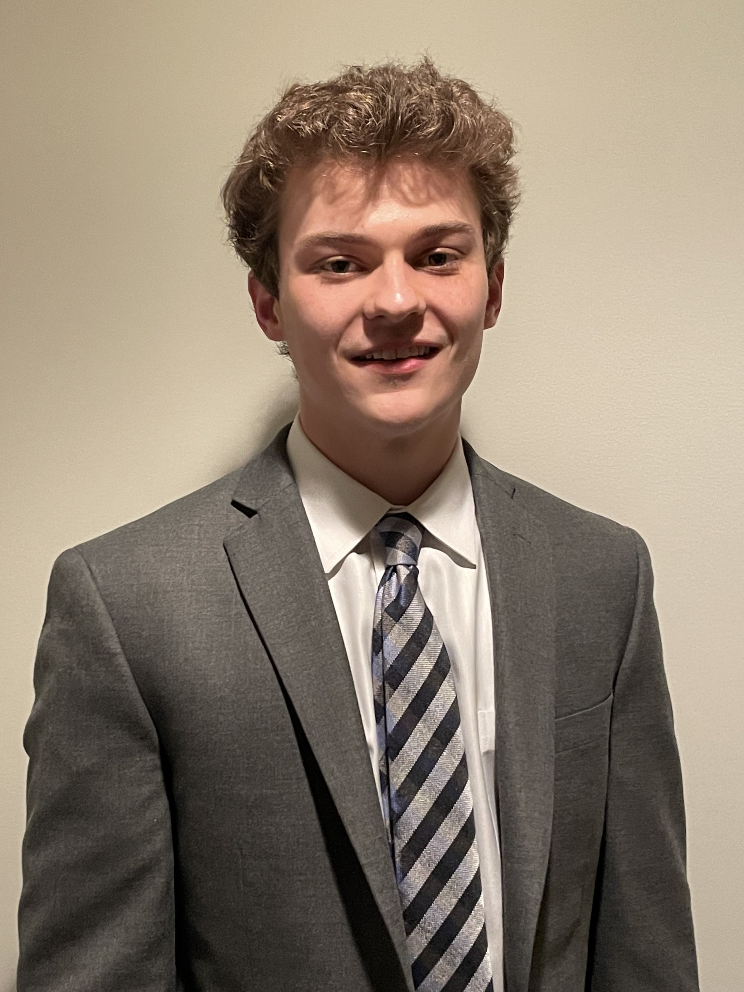 A young man in a gray suit, white shirt, and striped tie standing against a plain light-colored wall.