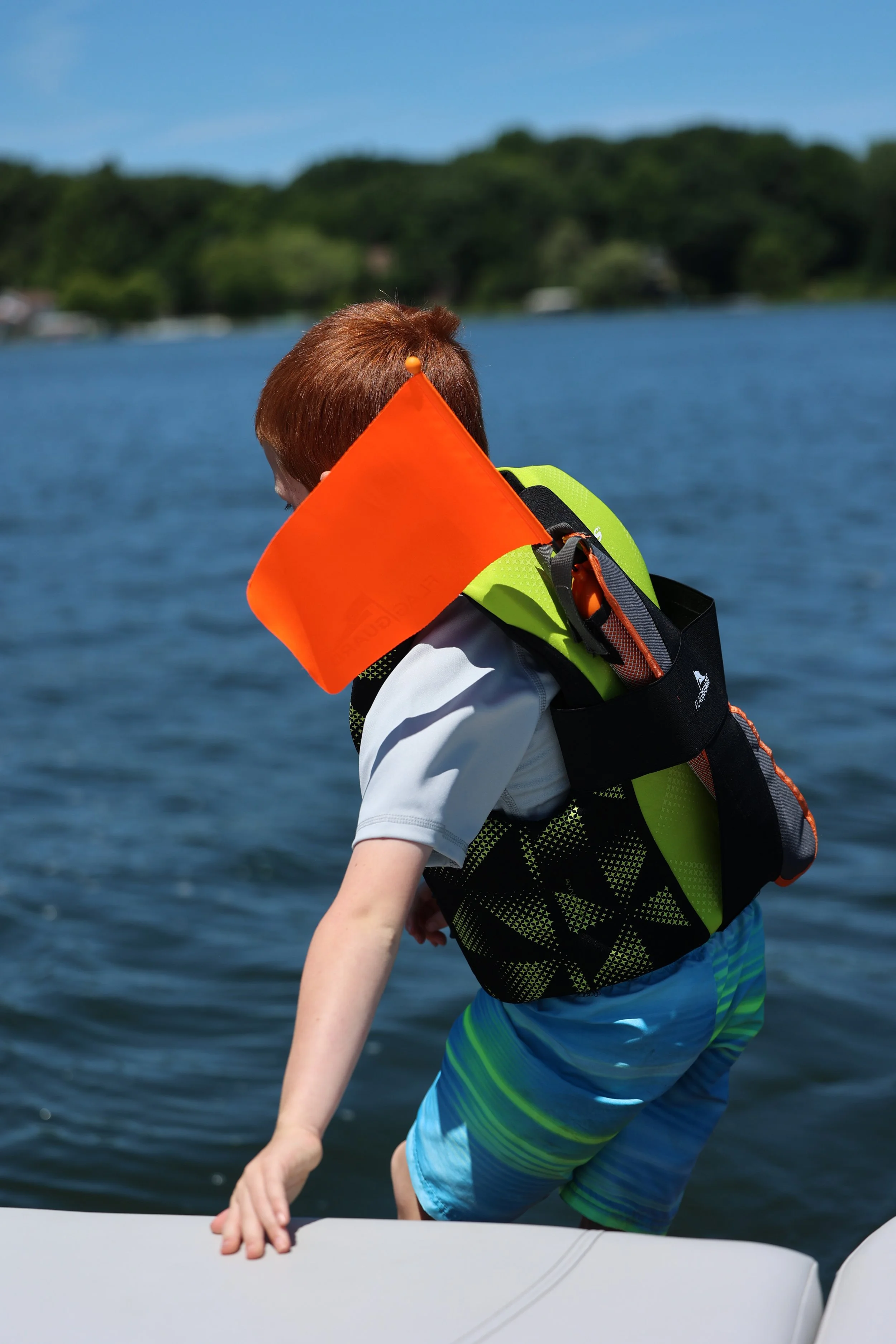 Child with red hair wearing a life jacket and colorful swim shorts reaches out with one hand on the edge of a boat on a lake, with trees and blue sky in the background.