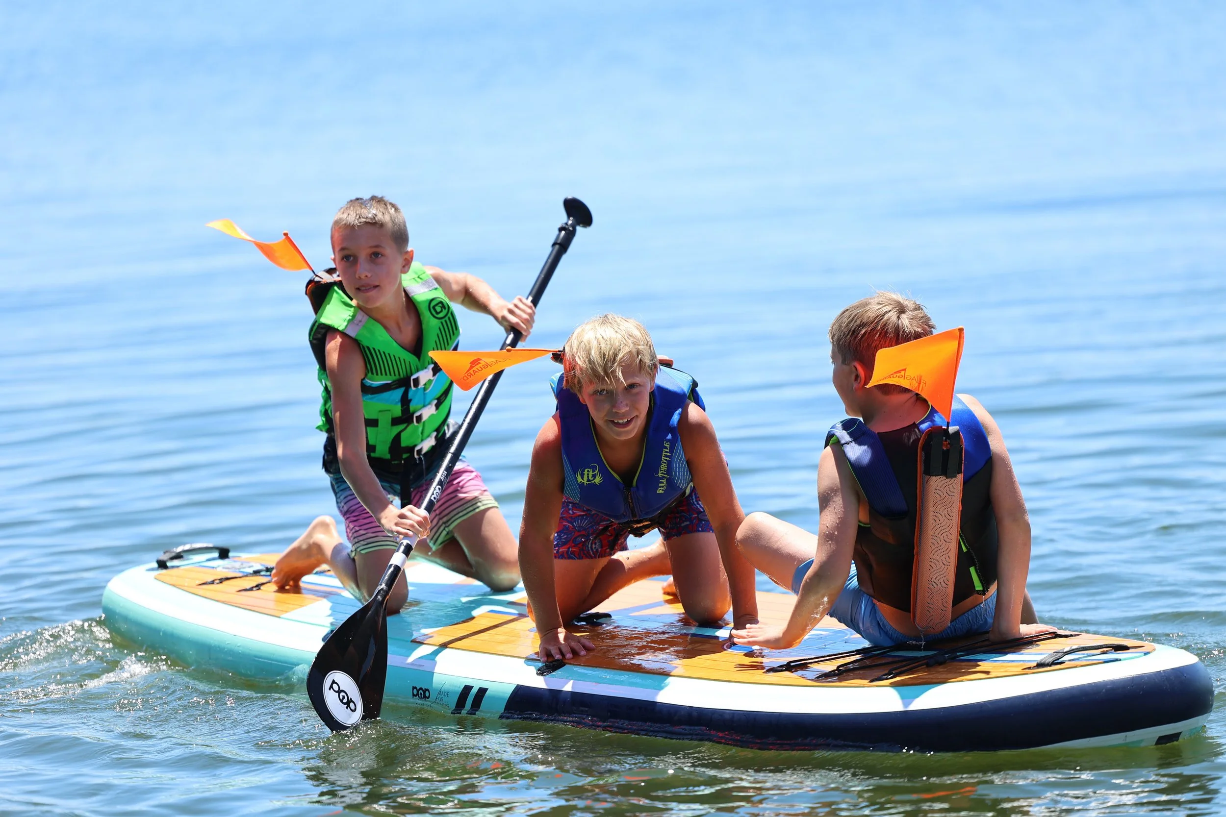 Four children on a paddleboard, three kneeling and one standing with a paddle in the water, wearing life jackets, on a sunny day