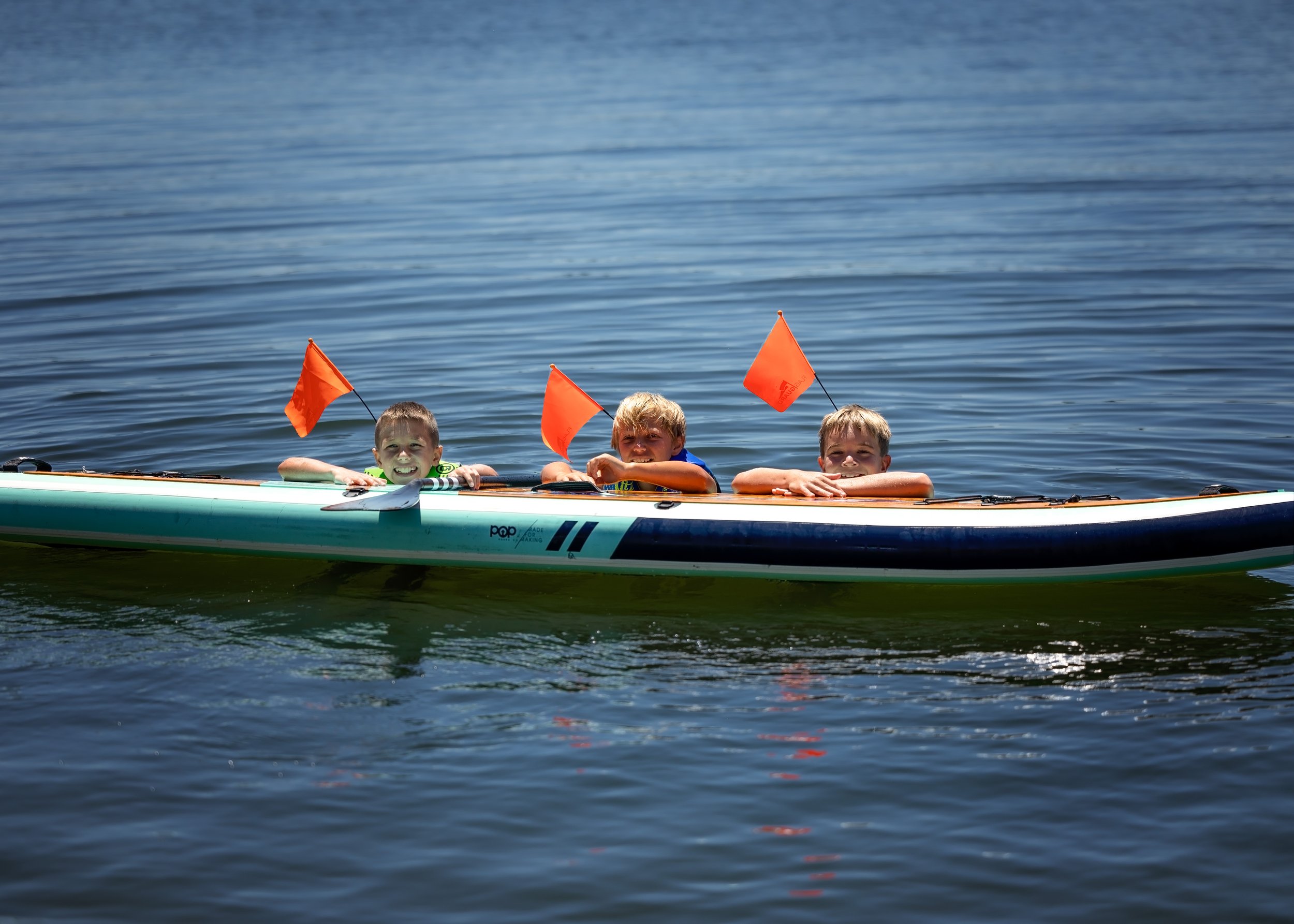 Three children holding orange flags and smiling while sitting in a paddle boat on a calm lake.