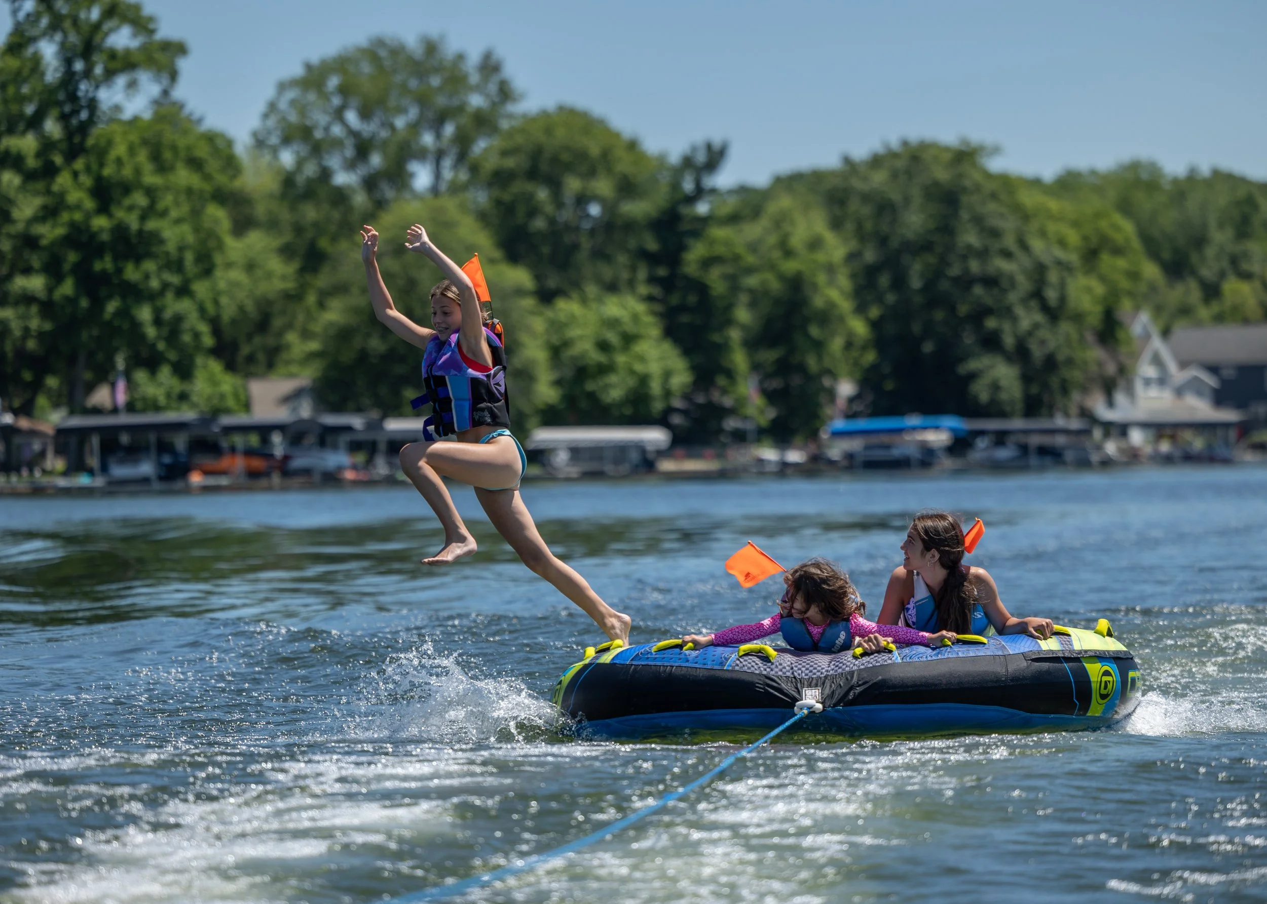 A group of young girls enjoying tubing on a lake, with one girl jumping off the tube into the water while others are seated on a floating platform, all wearing life jackets and floating gear, with trees and houses in the background on a sunny day.
