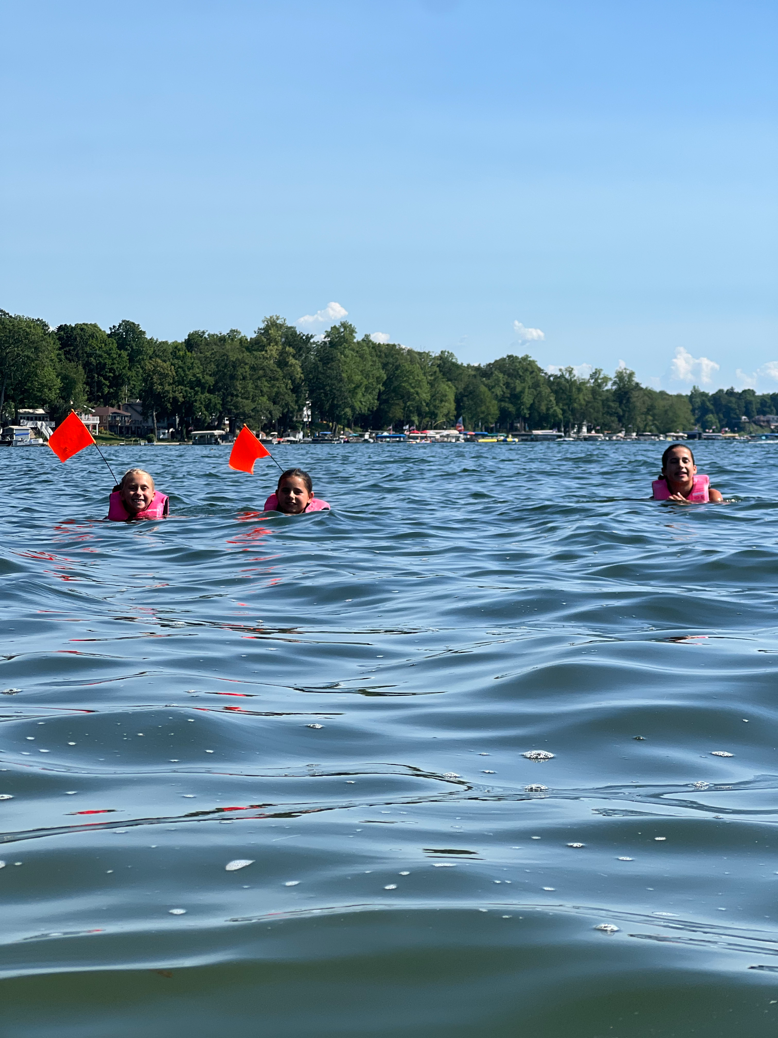 Three young women swimming in a lake with trees and houses in the background, wearing pink life jackets, with two of the girls using the patented FlagGuard system to increase visibility in the water.