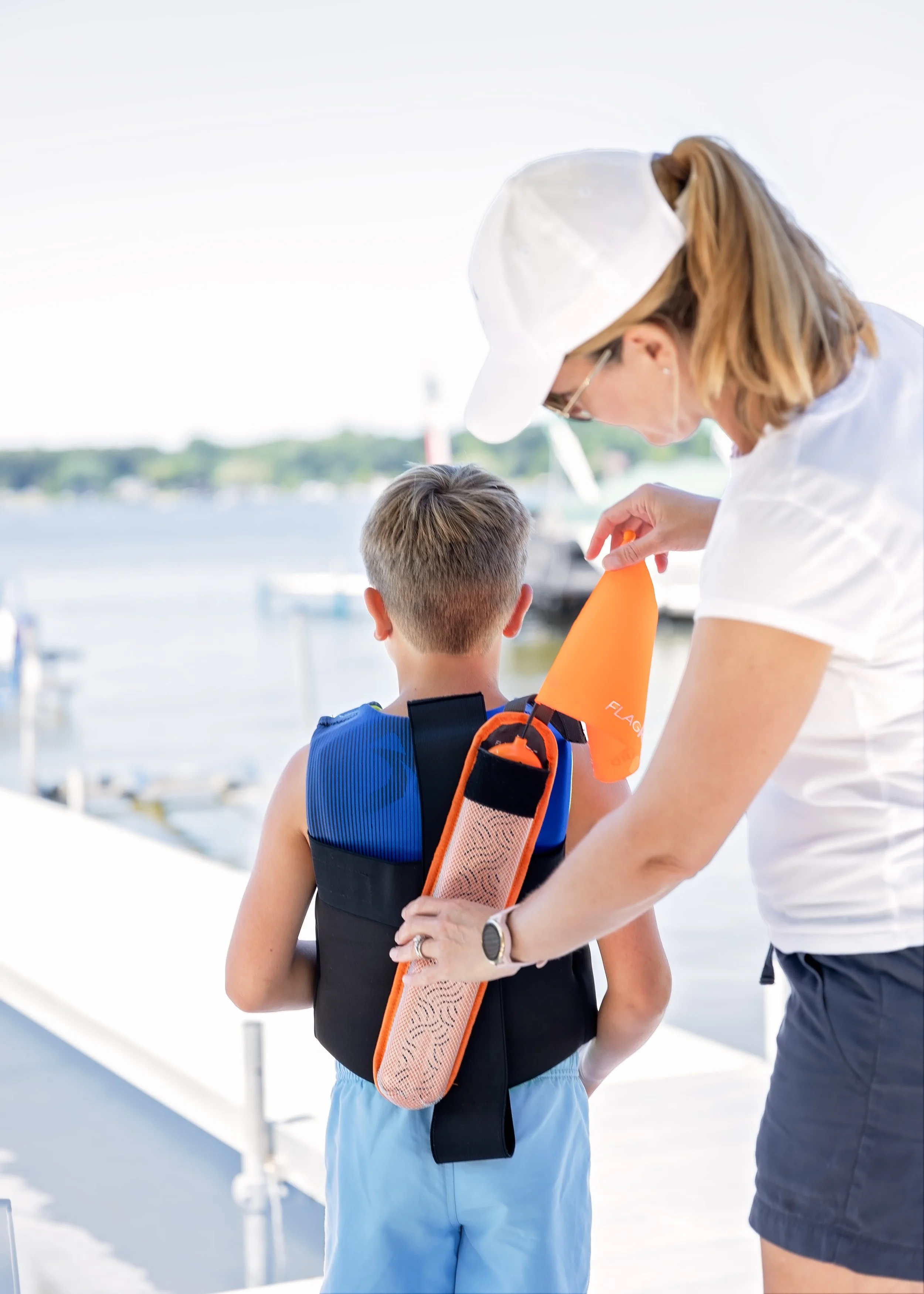 A woman is helping a young boy put on a life jacket and FlagGuard®, a patented water safety device, near a marina with boats and water in the background.