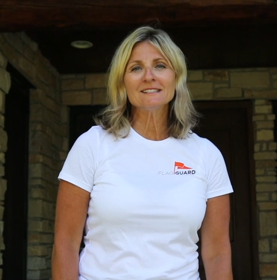 Founder and CEO, Erin McLaughlin, with blonde hair wearing a white T-shirt with a FlagGuard logo, stands outdoors in front of a stone wall and wooden structure.