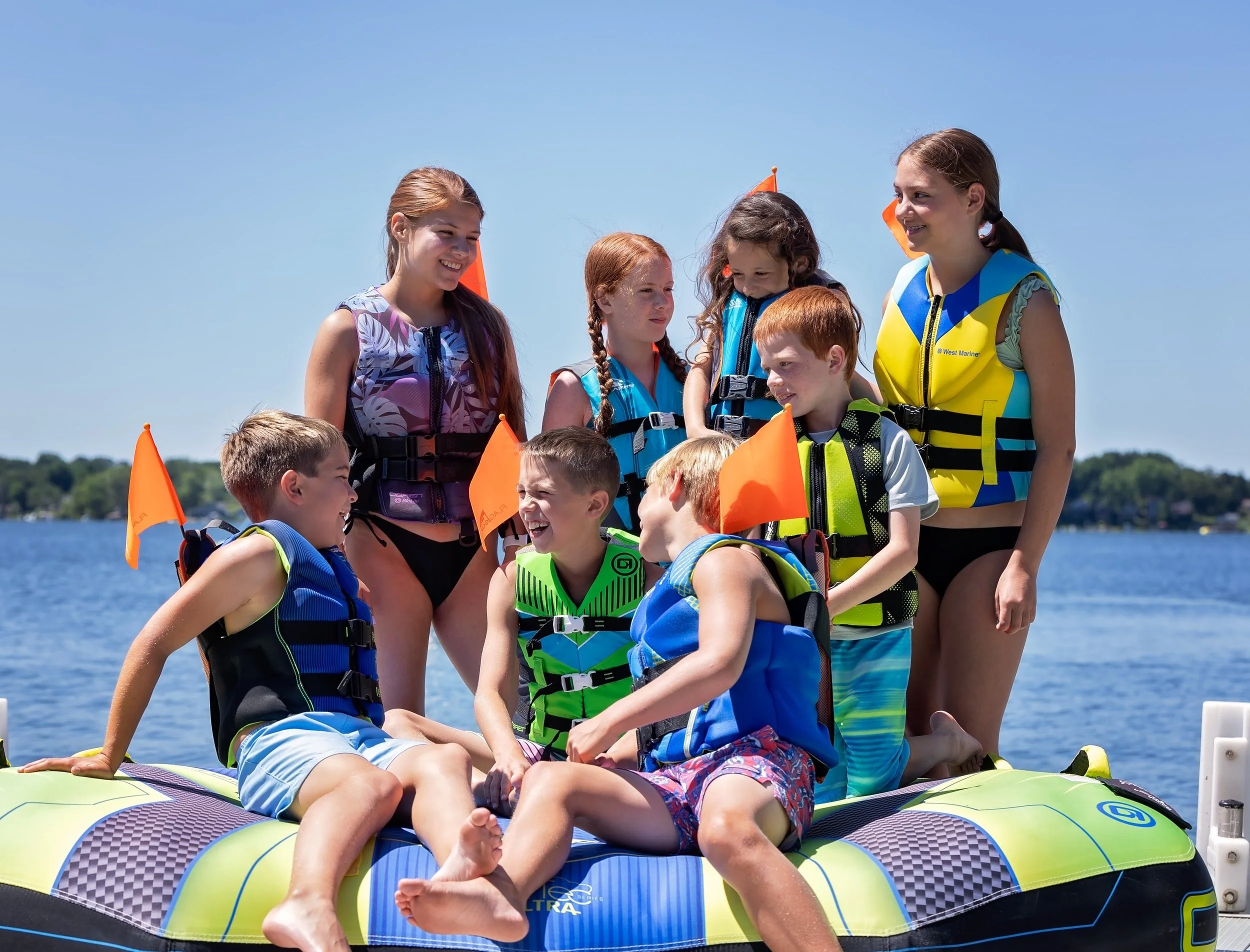 A group of children and two women on a boat at a lake, wearing life jackets and enjoying a sunny day.