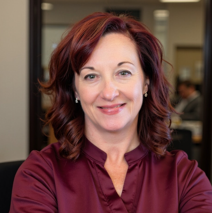 A woman with red hair, wearing a burgundy blouse, smiling in an office setting with a blurred background.