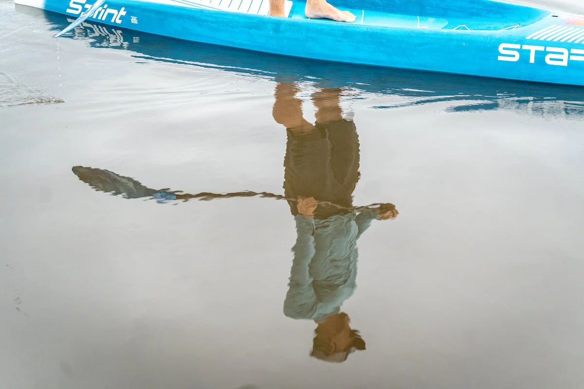 Person standing on the water's surface near a blue kayak or canoe, with their reflection visible in the water.
