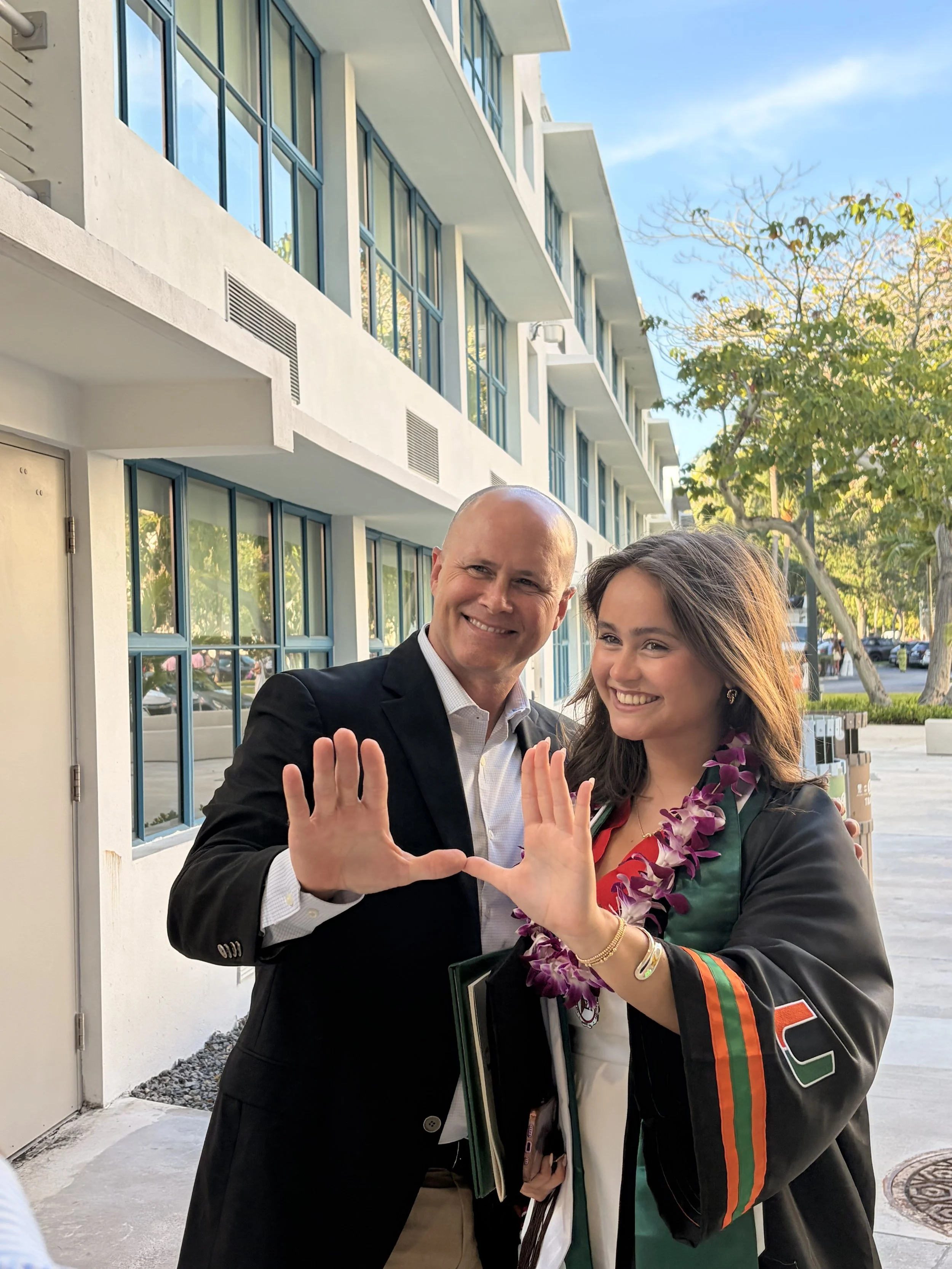 A man and woman smiling and making a heart shape with their hands. The woman is wearing a black jacket with orange and green stripes and a flower lei.