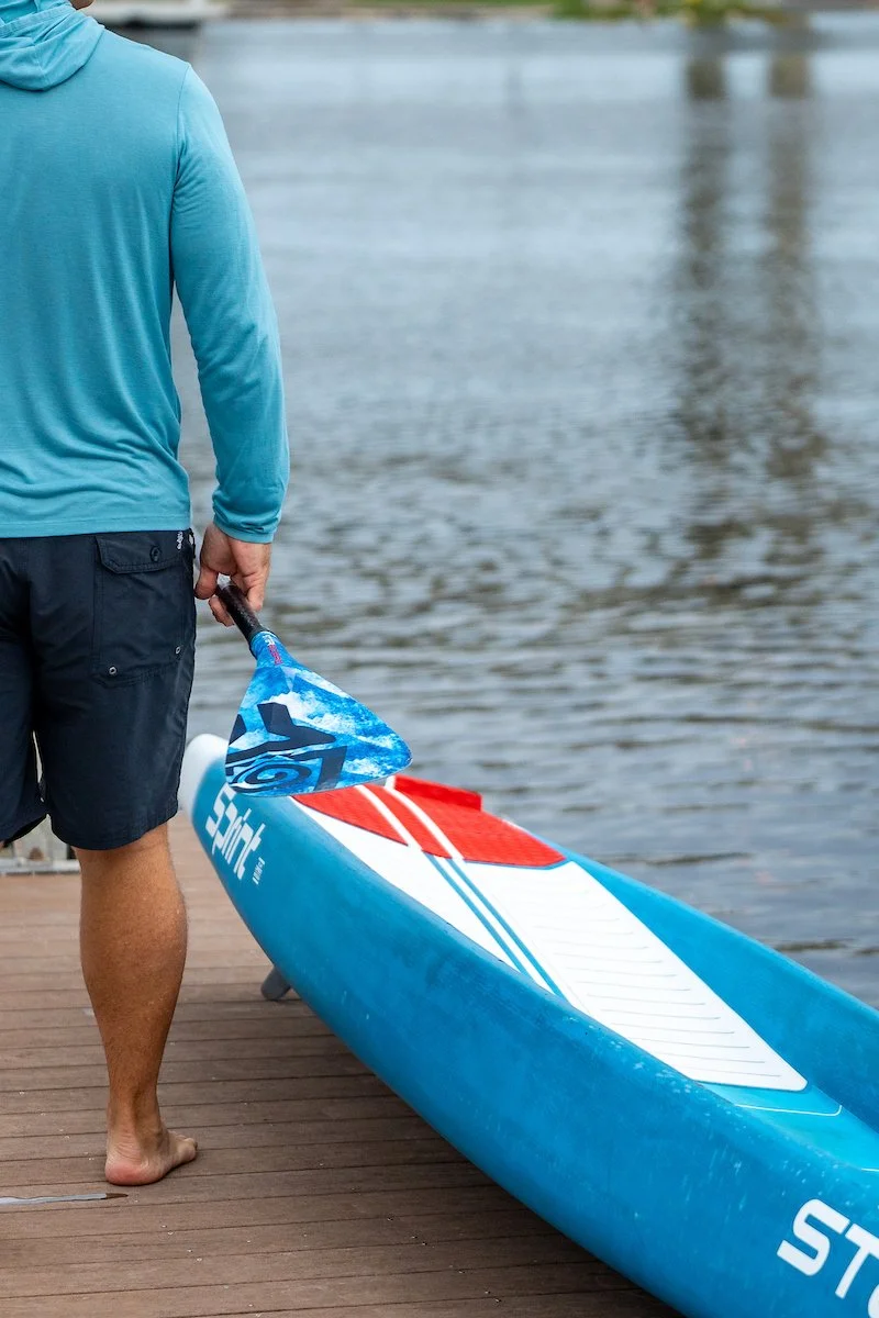 Person standing on a wooden dock holding a paddle next to a blue and white paddleboard, with water in the background.