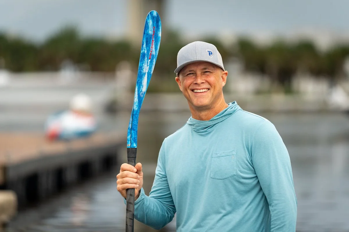 Smiling man in light blue shirt and gray cap holding a blue paddle near water with boats in the background.