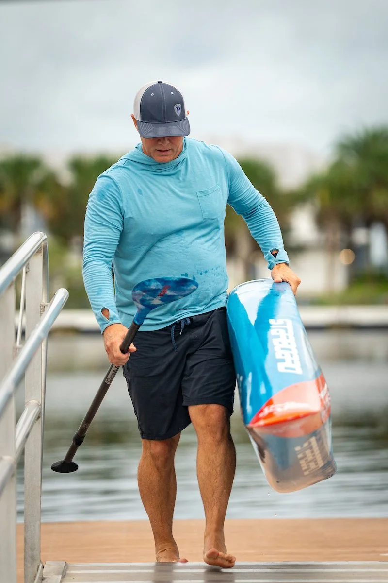 A man wearing a light blue long-sleeve shirt, black shorts, and a black and gray cap is holding a blue paddle and a paddleboard while walking barefoot onto a dock by the water, with palm trees in the background.