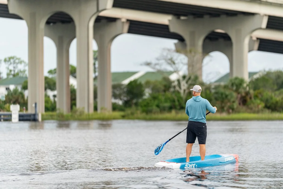 Man paddleboarding on a calm body of water under a large bridge, with greenery and trees in the background.