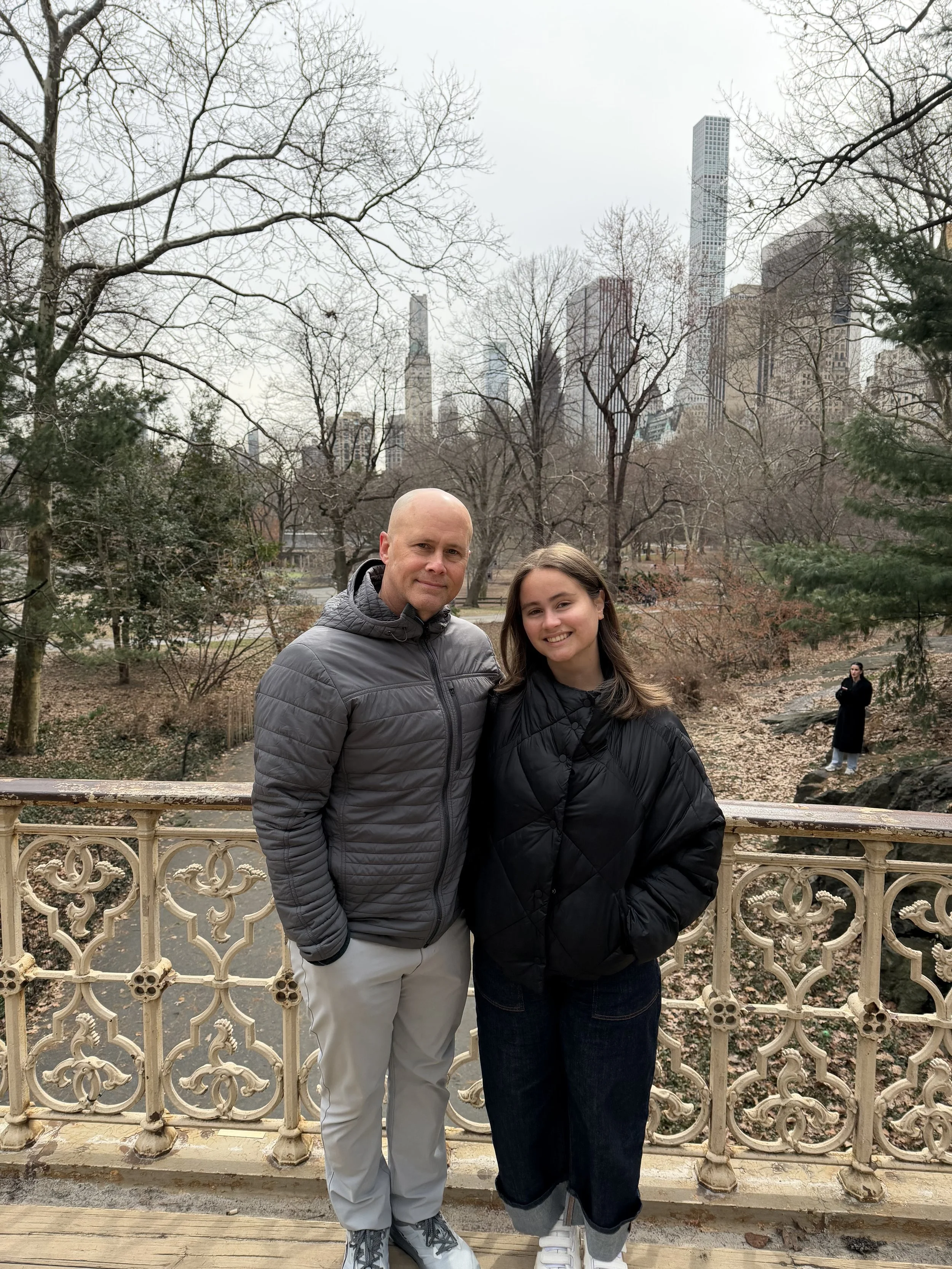 A man and a woman standing outdoors on a bridge with an ornate railing, surrounded by leafless trees and city skyscrapers in the background. The man is bald and wearing a gray puffer jacket, while the woman has long brown hair and is wearing a black puffer jacket.