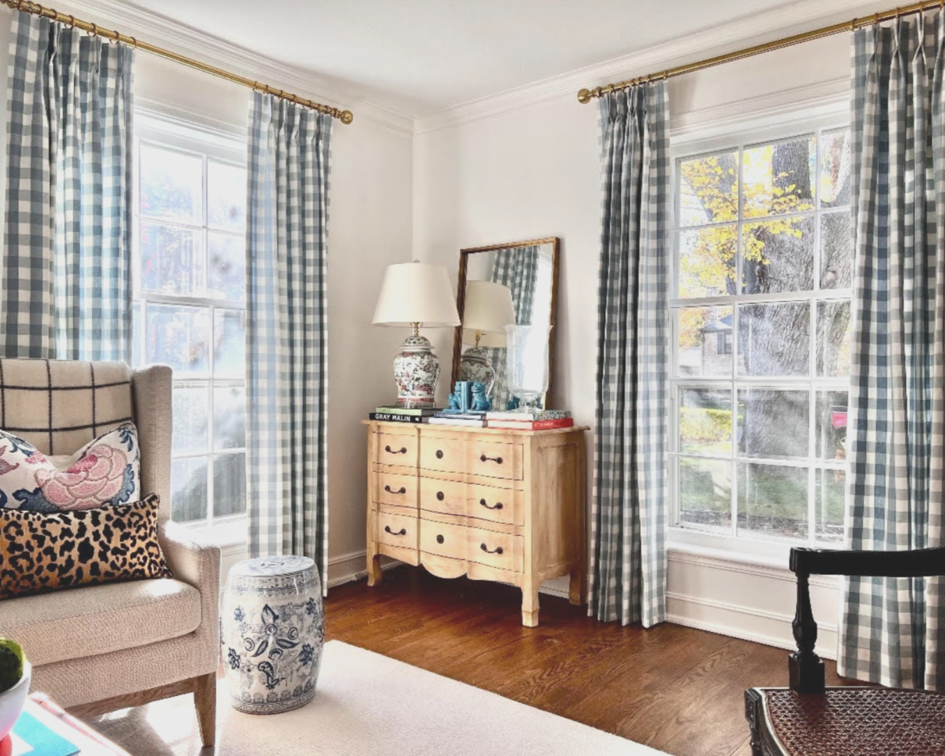 Living room with large windows, blue and white gingham curtains, a beige armchair with patterned pillows, a ceramic stool, a light wood dresser with a mirror and lamp, and hardwood floors.