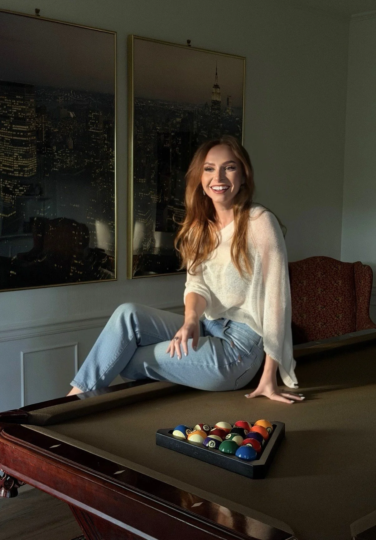 A woman with long auburn hair smiling and sitting on a pool table with her legs crossed and one arm resting on the table. Behind her, a wall features two framed pictures of cityscapes, with one prominently showing the New York City skyline including the Empire State Building. In the foreground on the pool table, there is a set of billiard balls arranged in a triangle rack.