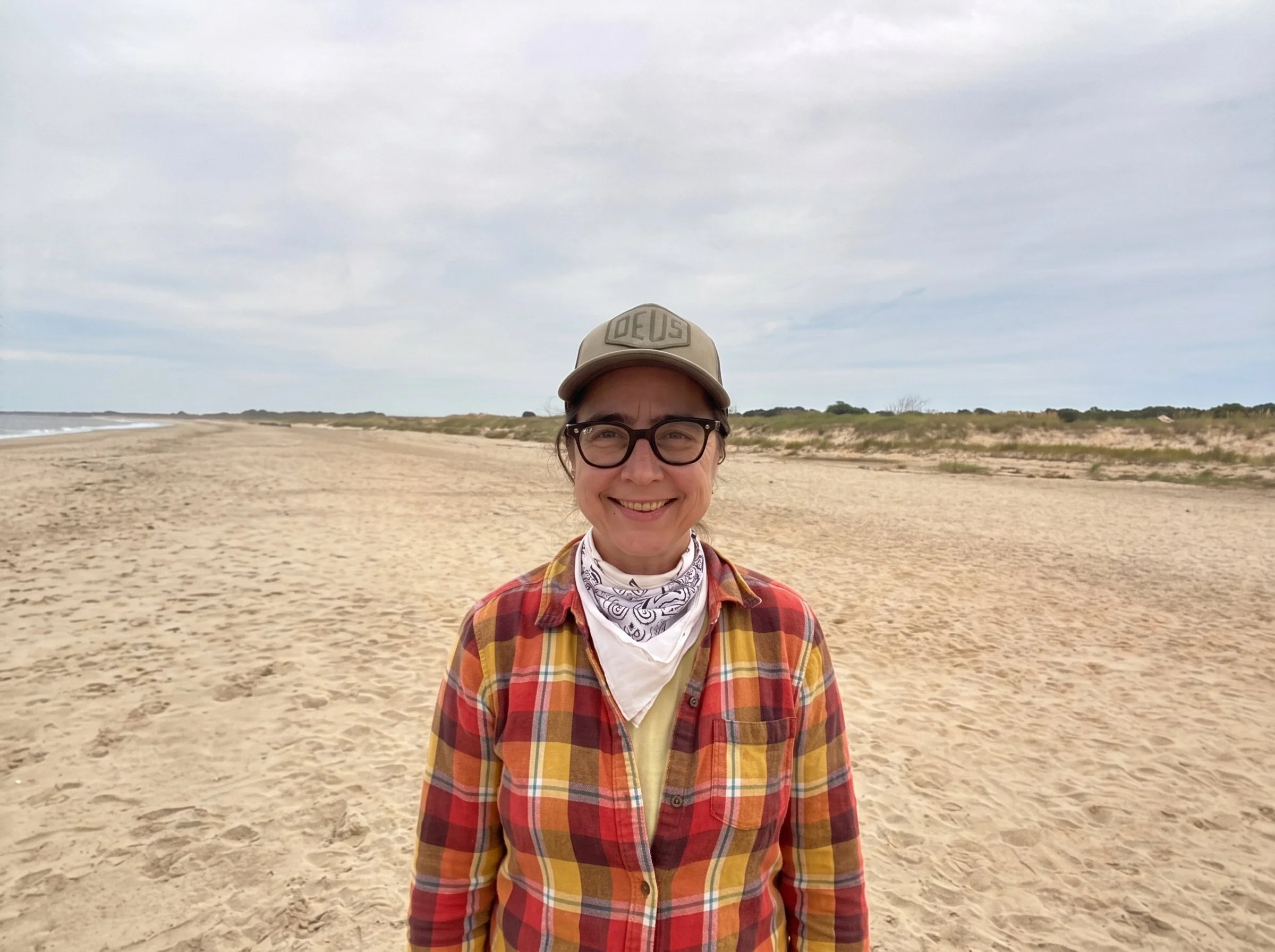 A smiling woman with glasses wearing a beige cap with the word 'Deus' on it, a white bandana around her neck, and a red and yellow plaid shirt. She is standing on a sandy beach with dunes and grassy areas in the background under a cloudy sky.