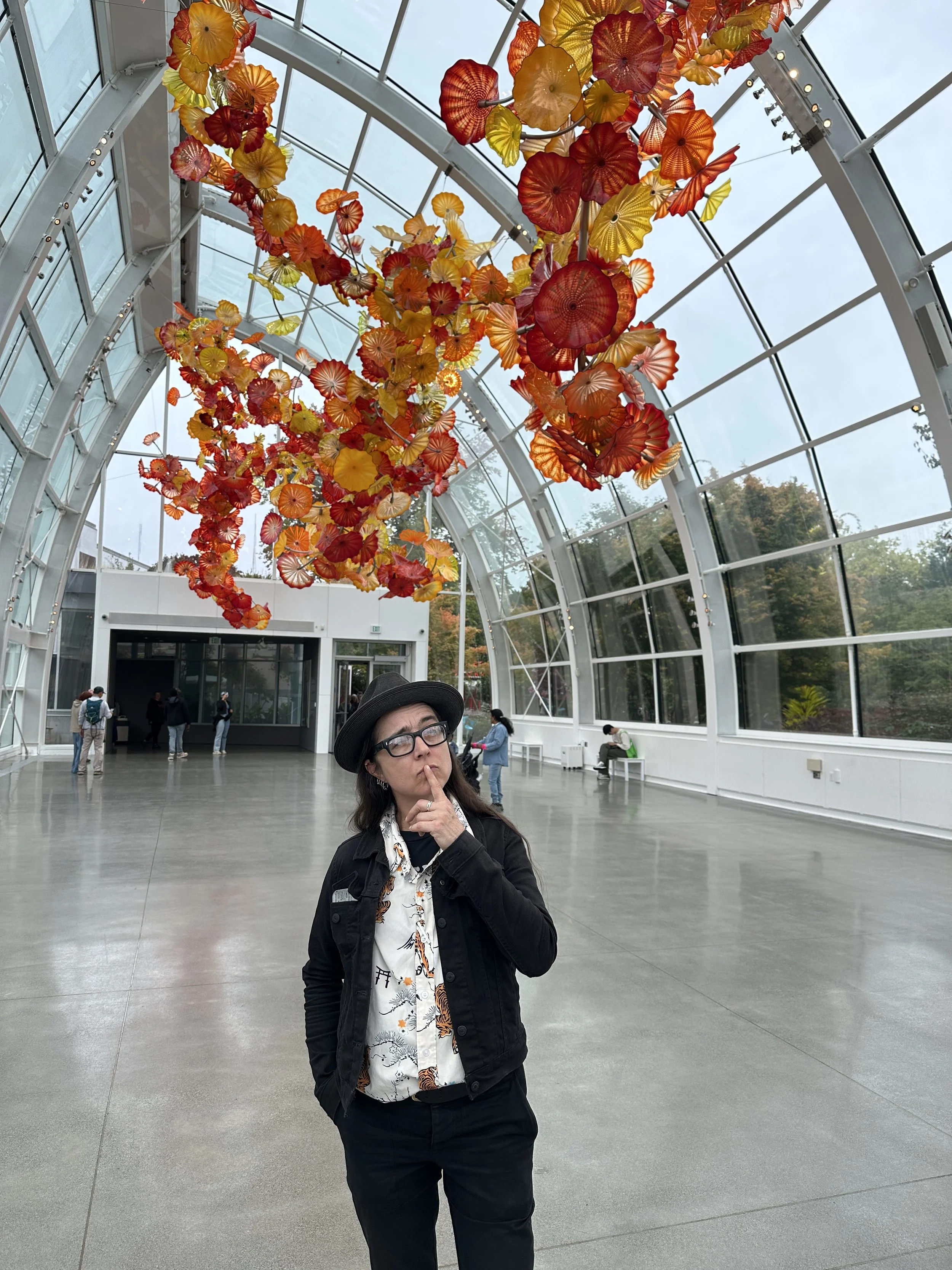 A woman with glasses, a black jacket, and a patterned shirt is standing inside a modern glass building with a curved ceiling. She is making a shushing gesture with her finger to her lips. Hanging from the ceiling are colorful glass sculptures resembling flowers in red, orange, and yellow shades. In the background, other people are visible, some sitting and some standing near a doorway.