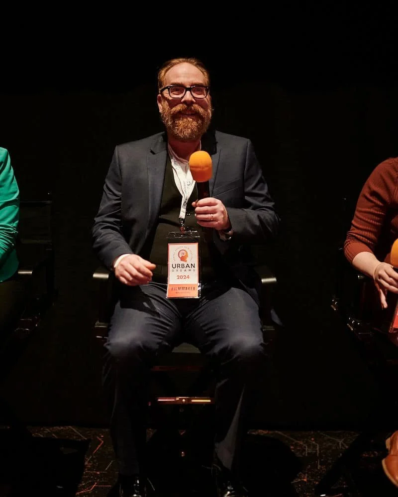 A man with glasses and a beard sitting on a chair, holding an orange microphone, wearing a suit jacket, white shirt, and a lanyard with a badge reading "URBAN FILMMAKER 2024."