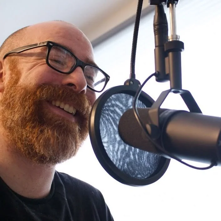 A man with glasses and a red beard smiling in front of a professional microphone setup, possibly recording audio or podcasting.