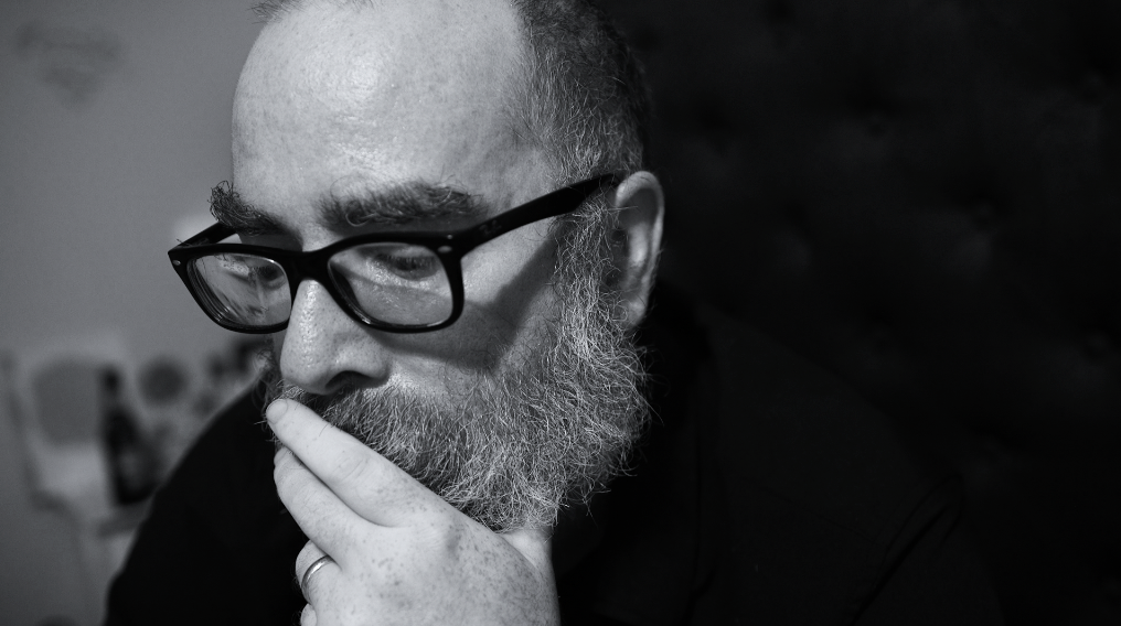 Black and white close-up of a man with glasses, beard, and bald head, holding his chin thoughtfully.