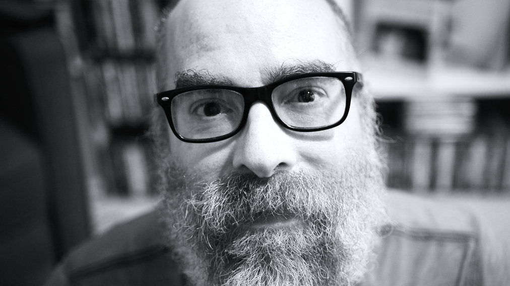 A close-up black and white photo of a man with glasses and a full beard, looking directly at the camera. Behind him are bookshelves.