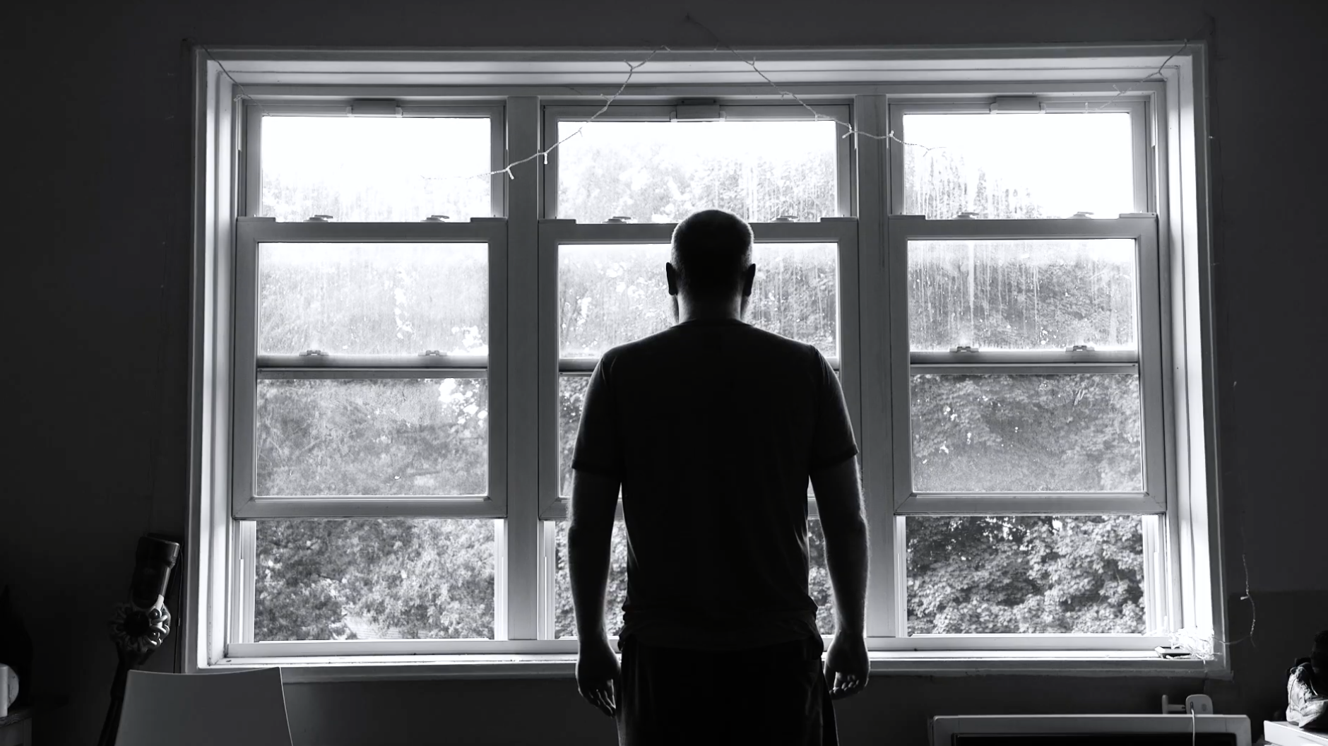 A person with short hair standing indoors, looking out of a large window with four panes, revealing trees outside. The image is in black and white.