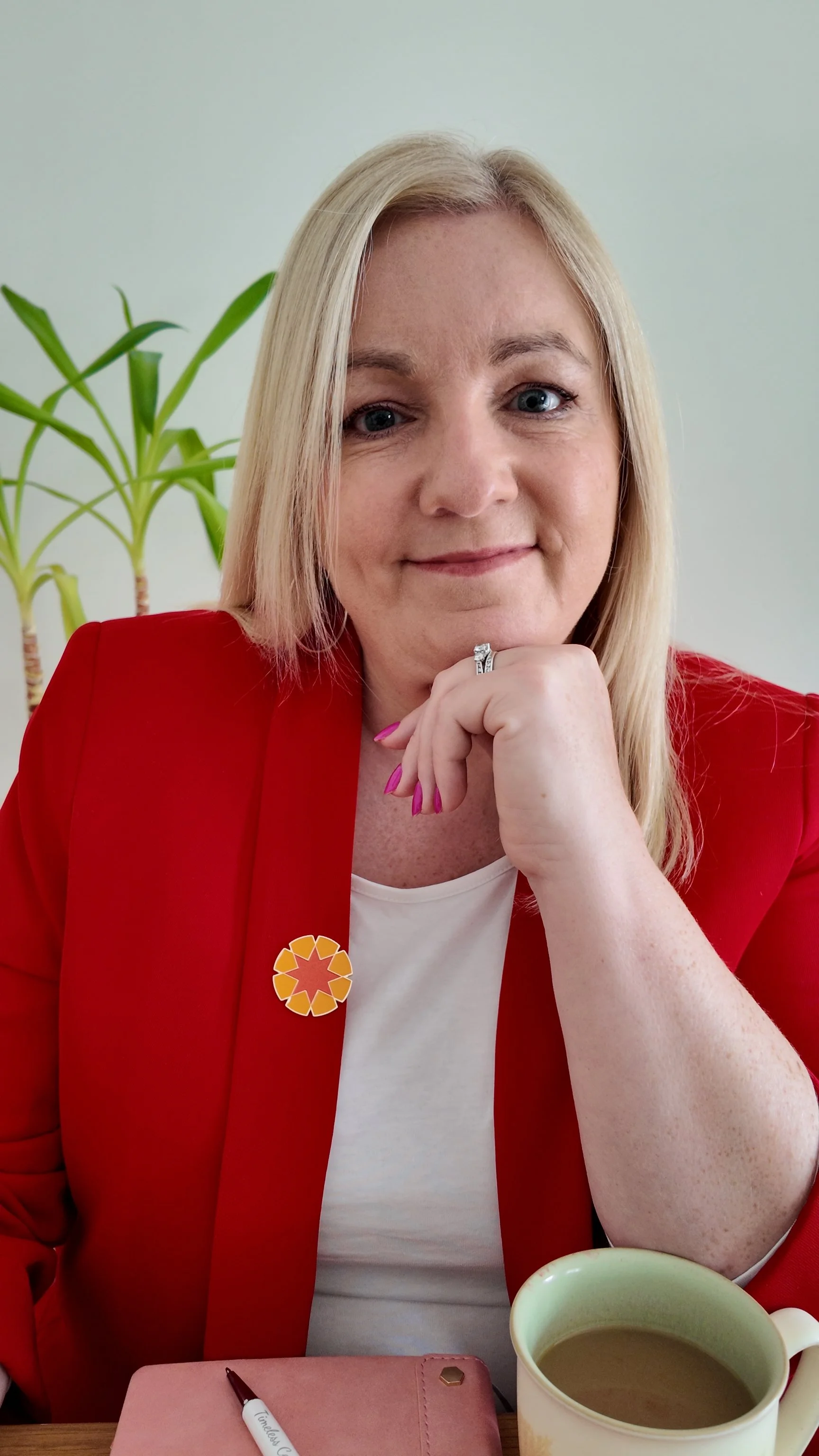 A woman with blonde hair wearing a red blazer and white top sitting at a table with a mug of tea, a pink notebook, and a pen, smiling with her hand resting on her chin, in front of a light green wall and a green plant in the background.