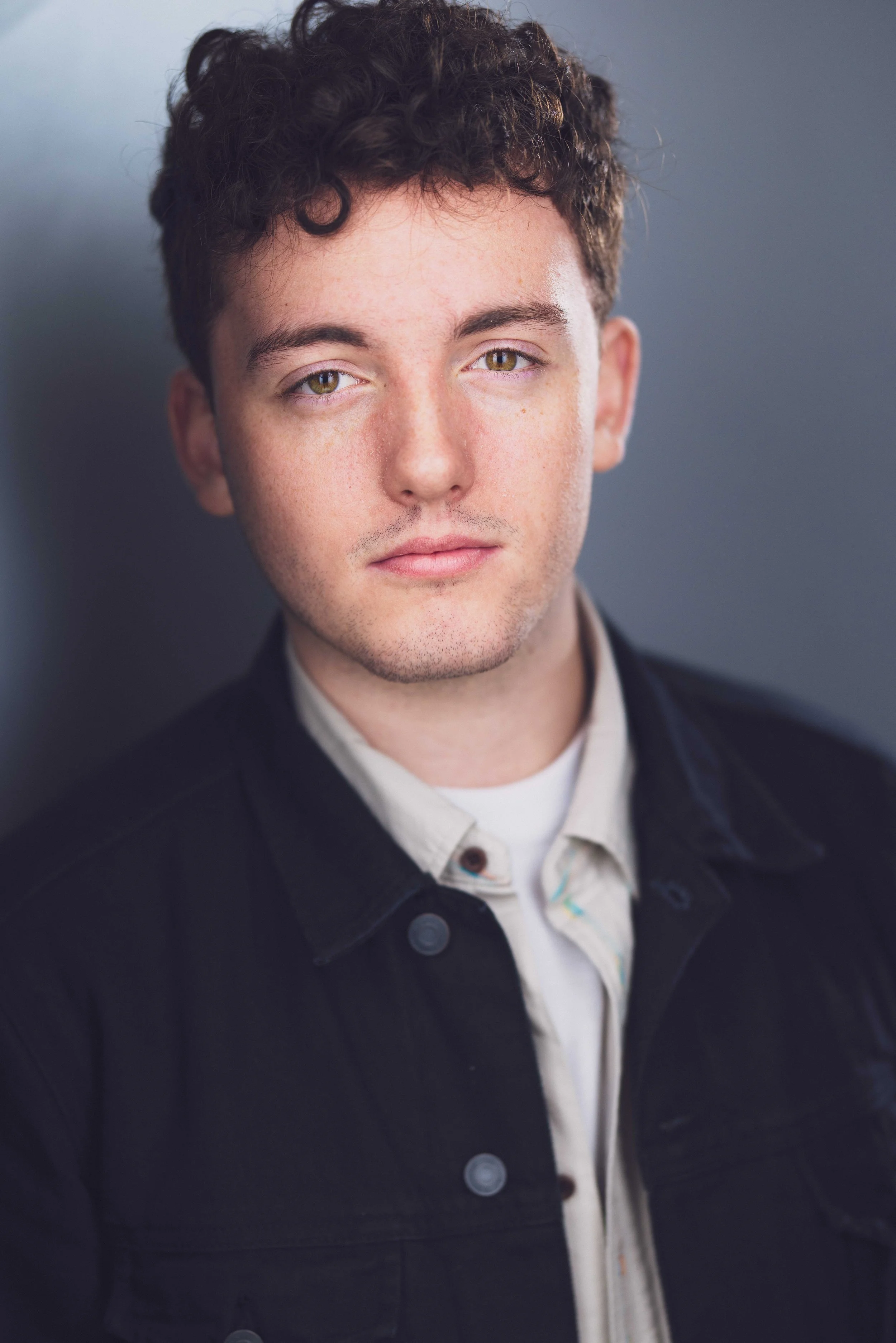 Close-up of a young man with short curly brown hair, hazel eyes, and light skin, wearing a white shirt and a black jacket, looking directly at the camera against a gray background.