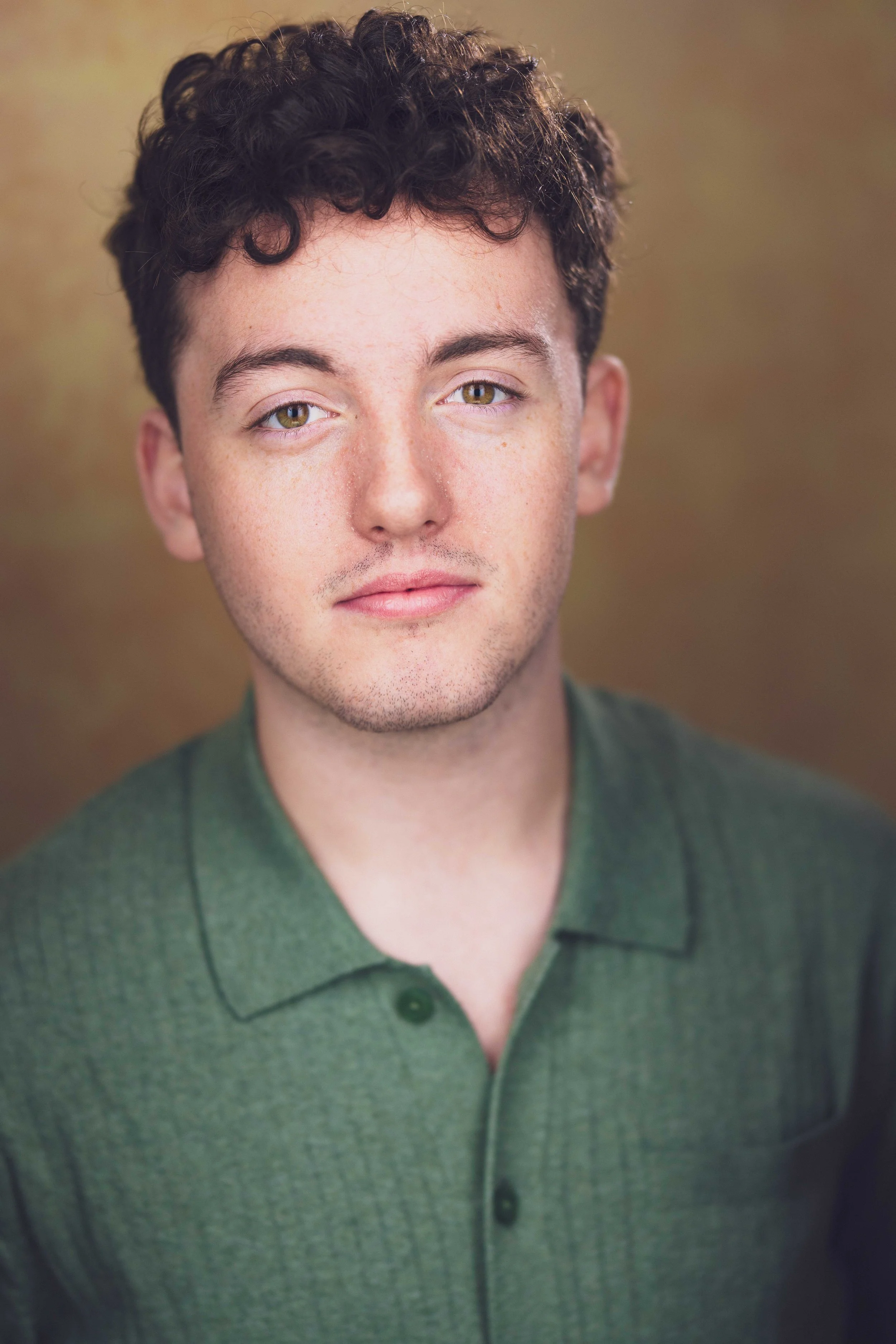 A young man with short curly dark hair, hazel eyes, and light skin wearing a green collared shirt.