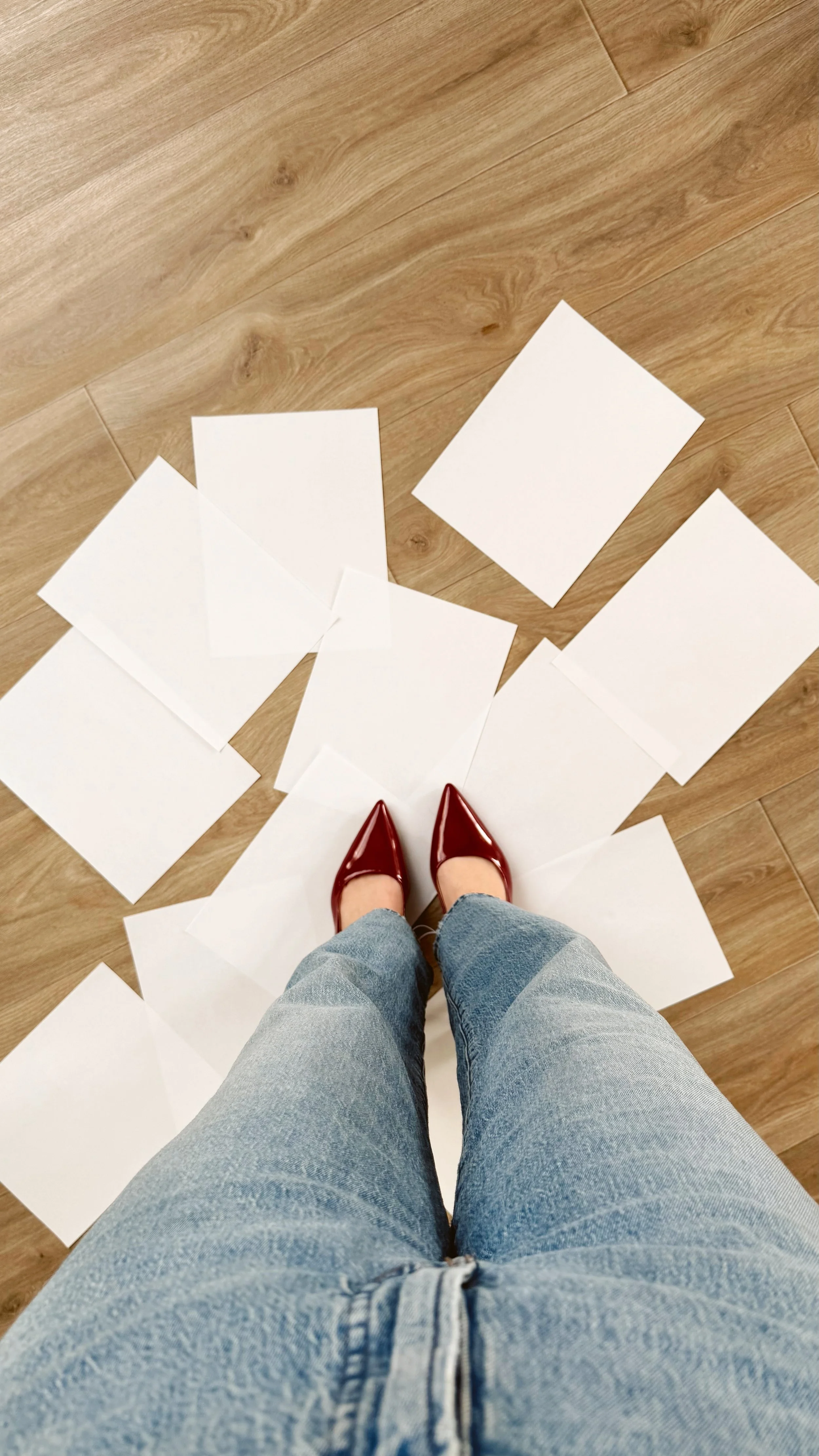 Person standing on wooden floor with scattered blank white papers and wearing red shiny shoes and blue jeans.