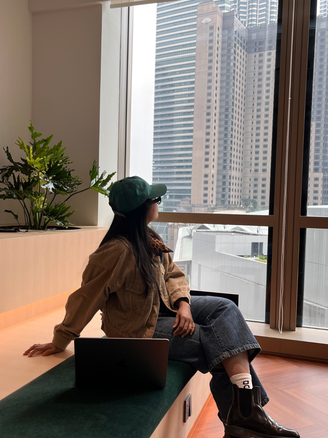 A young woman sitting on a green bench near a large window with a cityscape view, wearing a green cap, glasses, a brown jacket, a black skirt, white socks, and black boots, with a laptop beside her.