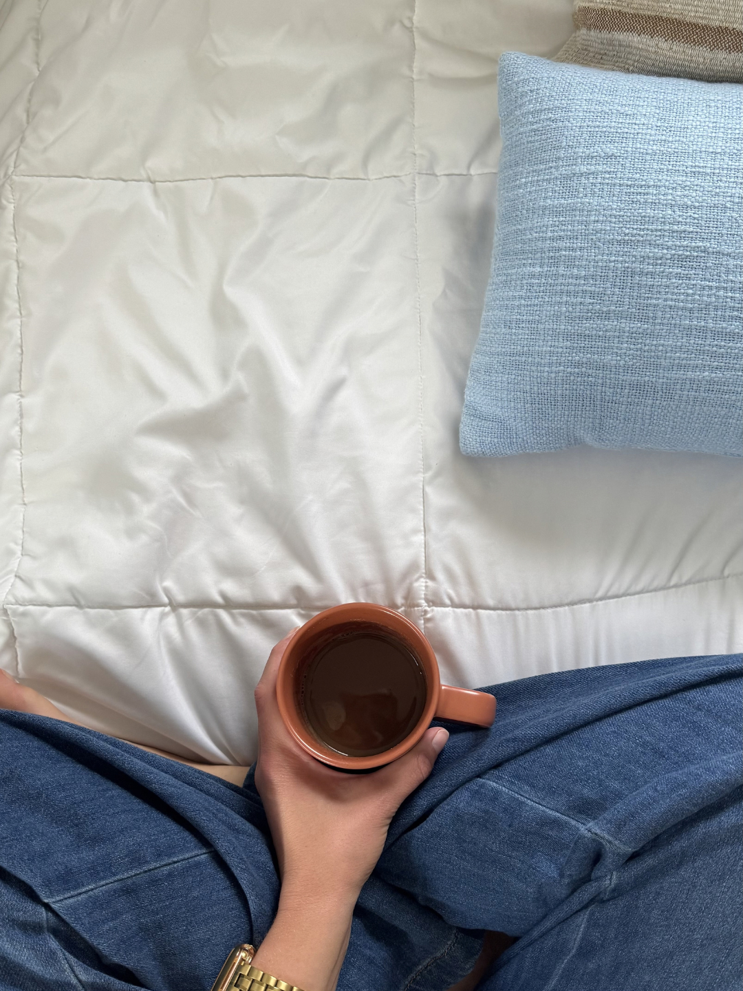Person sitting on a bed with a cup of coffee, wearing blue jeans and a gold watch; the bed has a white comforter and a light blue pillow.