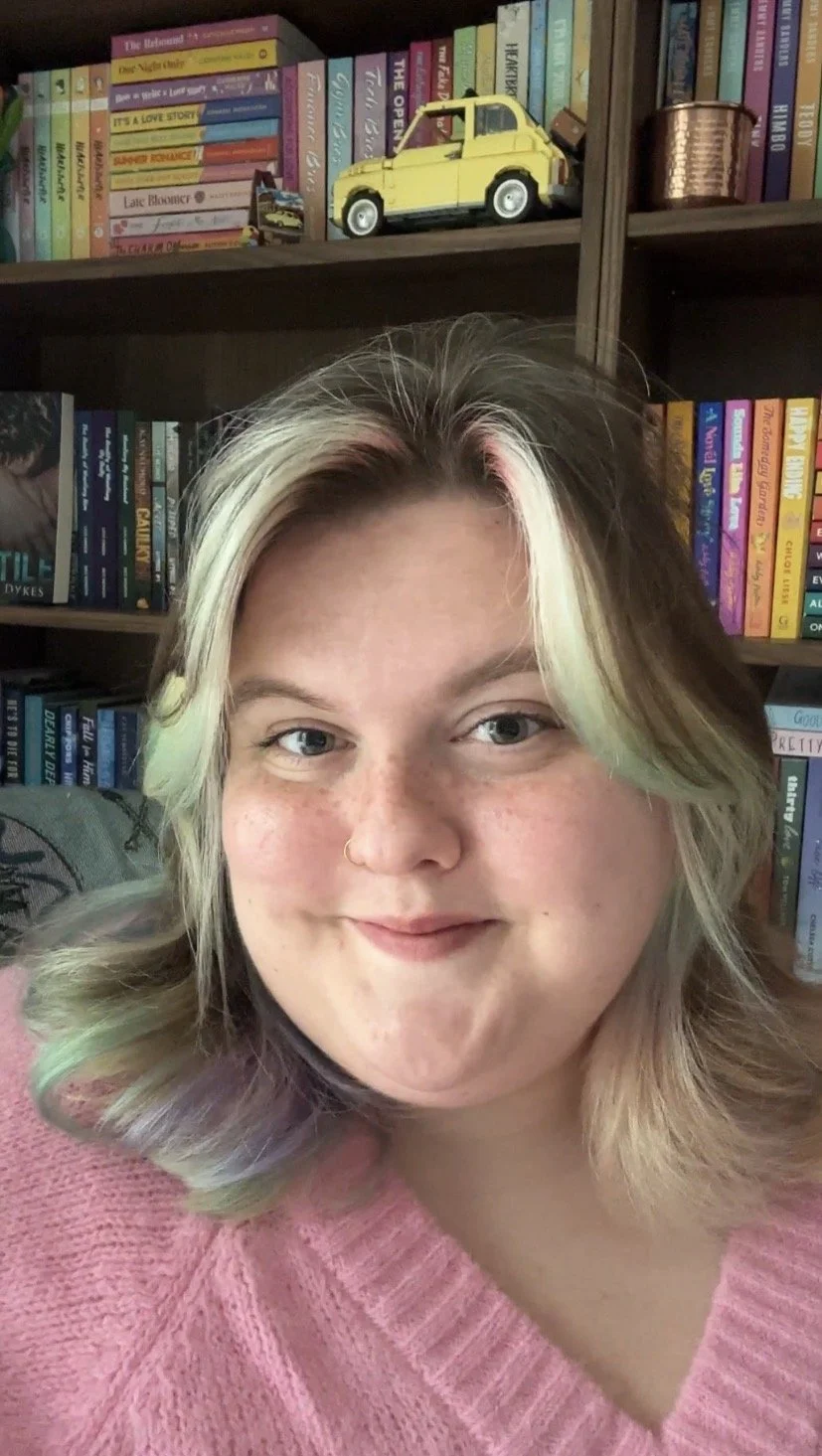 Young woman with blonde hair and a pink sweater, smiling in front of a bookshelf filled with colorful books and decorative items, including a yellow toy truck.
