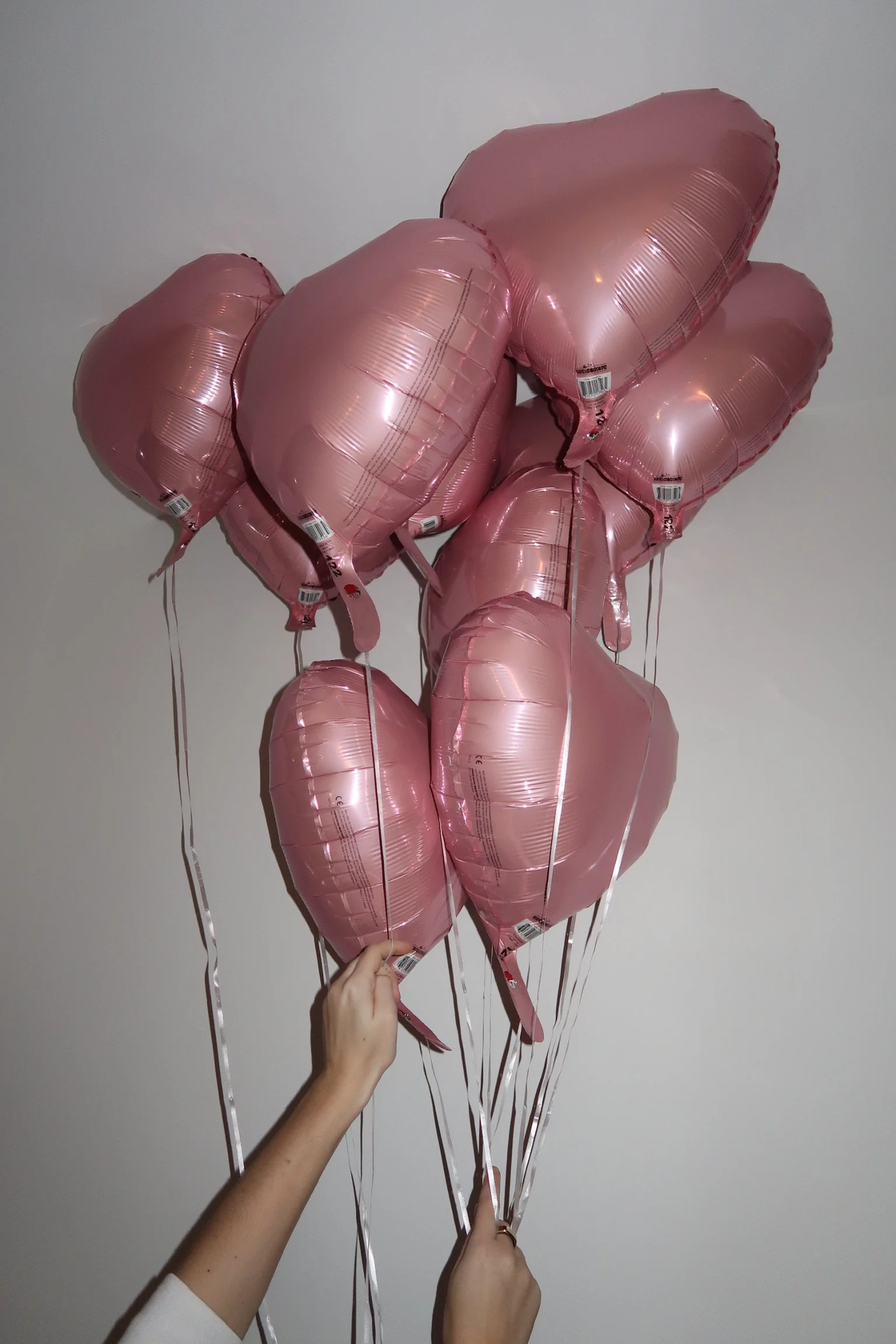 A group of pink heart-shaped foil balloons being held by a person with both hands, against a plain gray wall background.