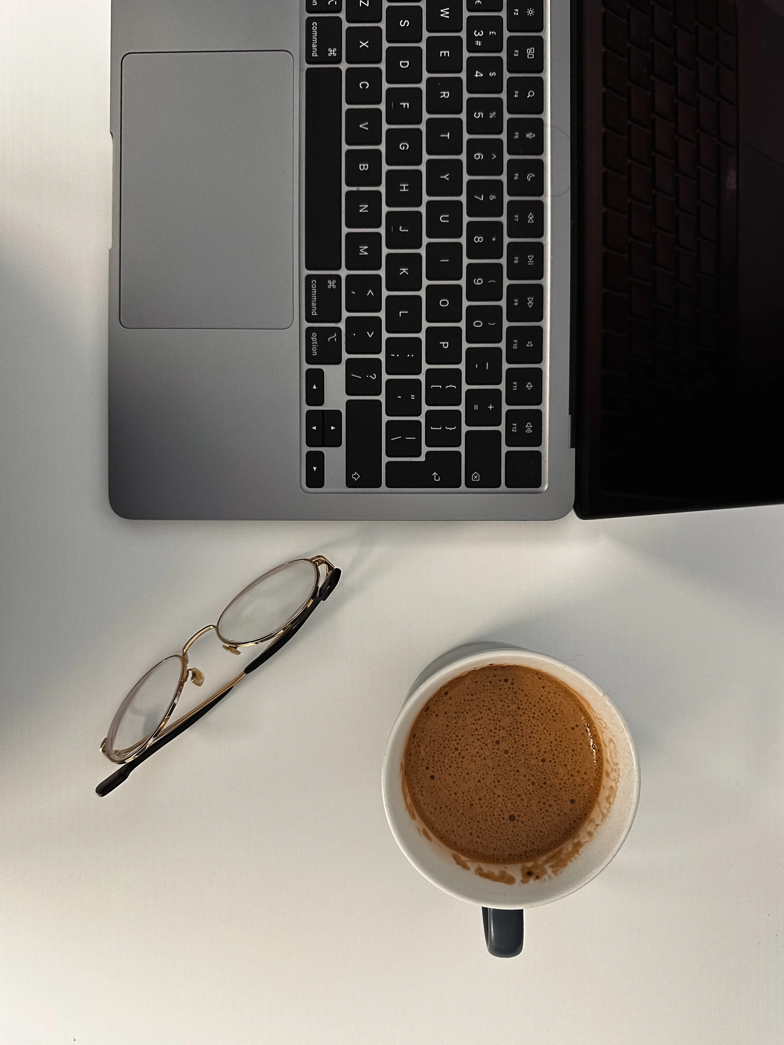 A laptop keyboard, eyeglasses, and a cup of coffee on a white table.