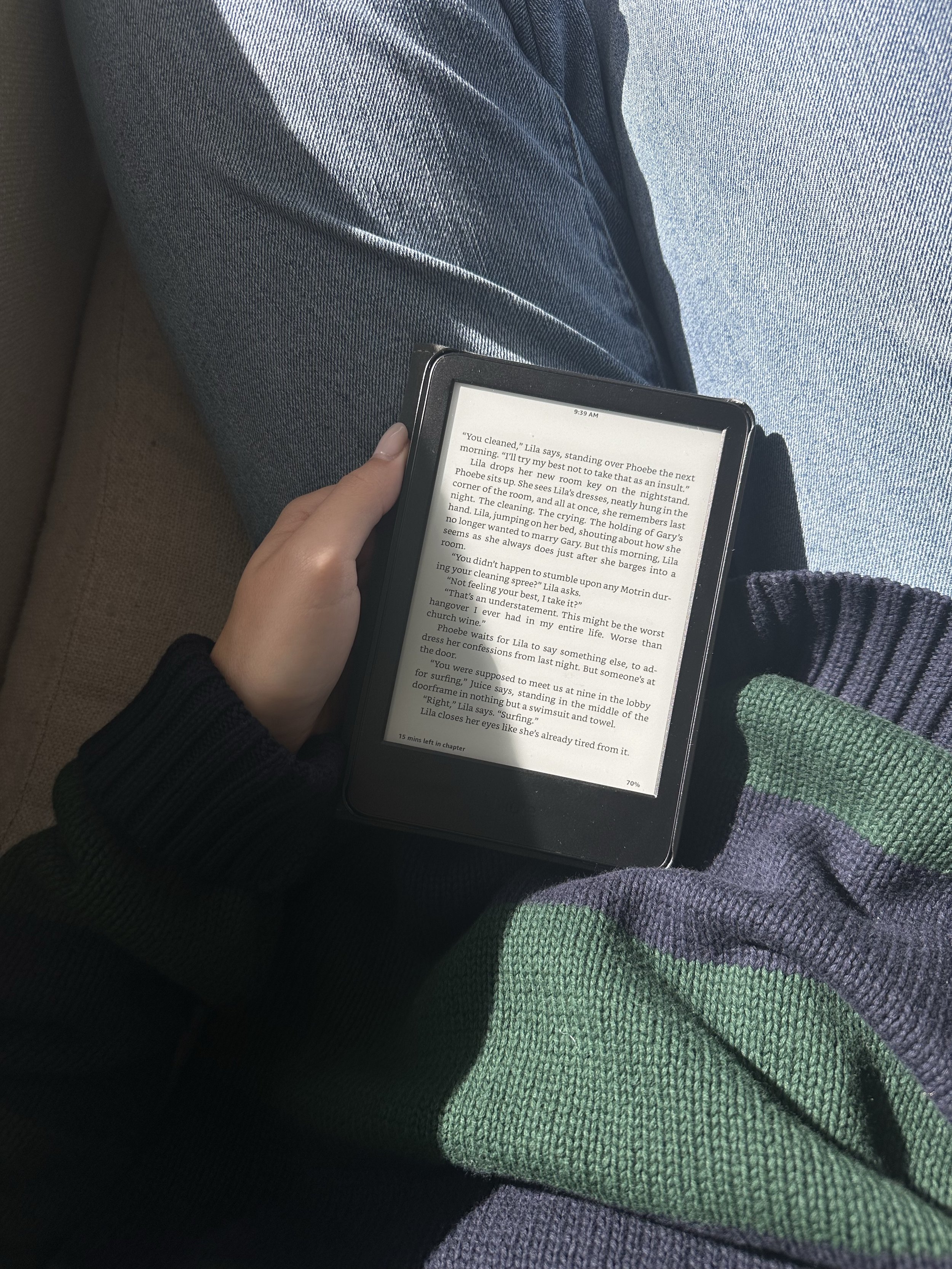 A person sitting on a dark seat, wearing blue jeans and a black sweater, holding an e-reader displaying a book interior, with a green and purple striped sweater sleeve partially visible.