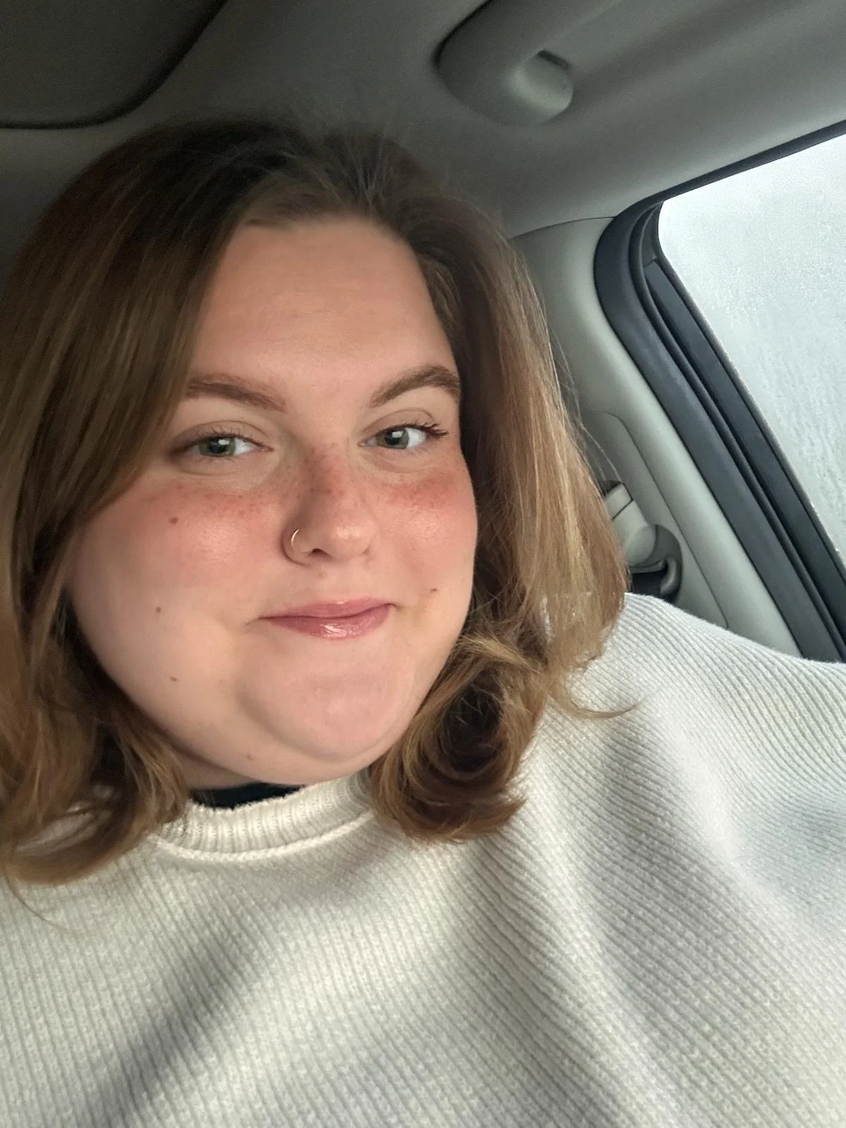 A woman with shoulder-length light brown hair and a nose ring is sitting in a car wearing a cream-colored sweater, smiling slightly at the camera.