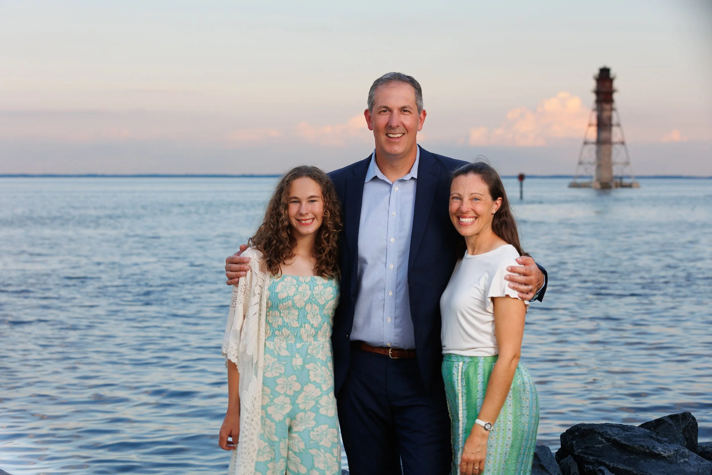 A family of three standing by the water with a lighthouse in the background during sunset or early evening. They are smiling and dressed in casual, colorful clothing.