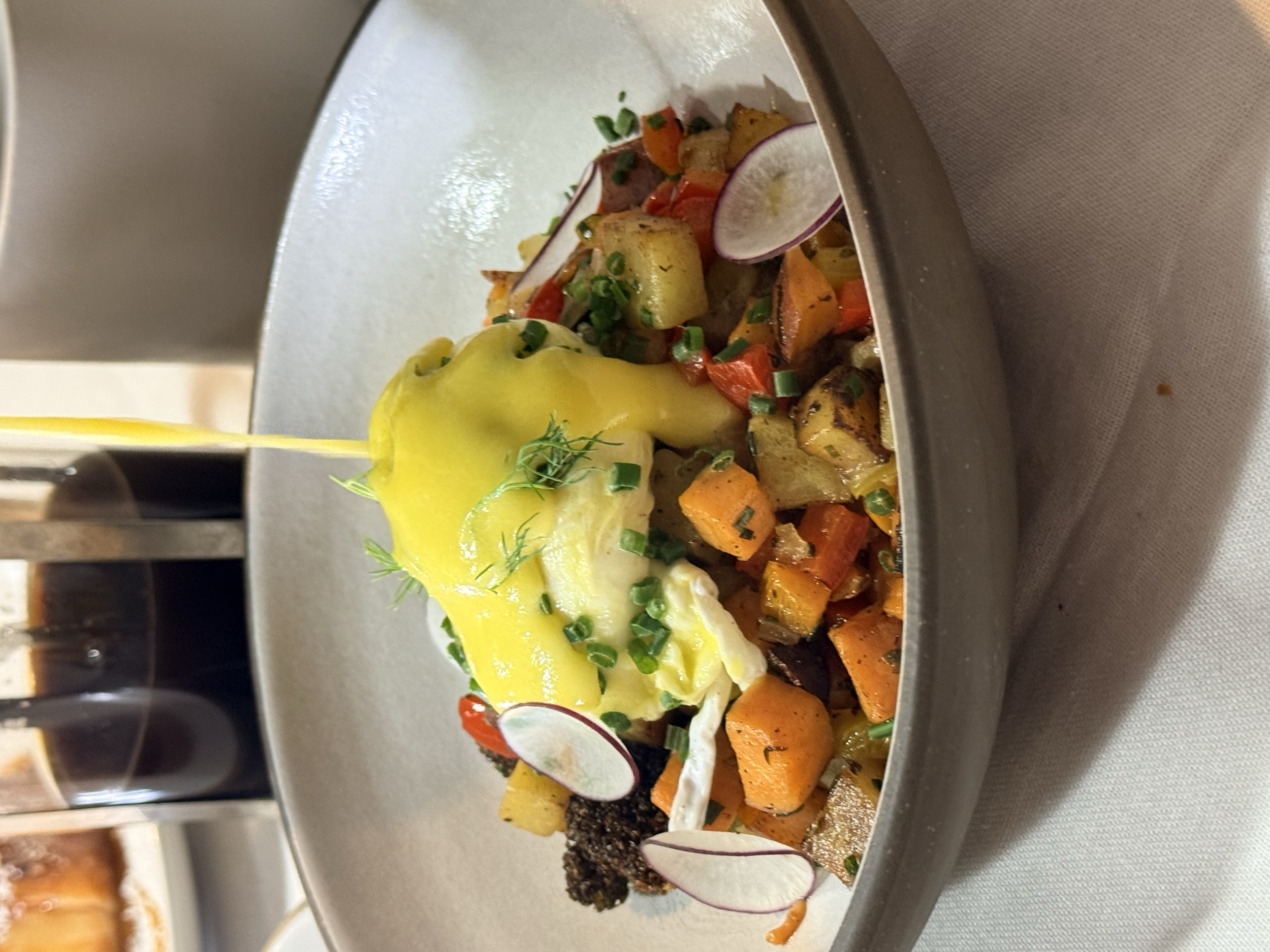 A bowl of roasted sweet potatoes topped with poached eggs, chopped chives, and microgreens, with yellow hollandaise sauce being poured over the eggs.